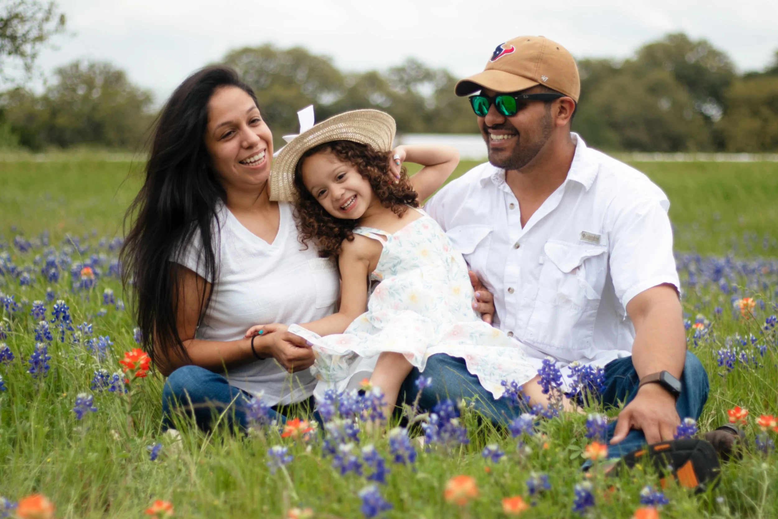 Family portrait by Levs Photography in Brenham, Texas featuring a happy family of three sitting in a field of bluebonnets and wildflowers, smiling together on a sunny spring day with soft natural light and vibrant seasonal colors