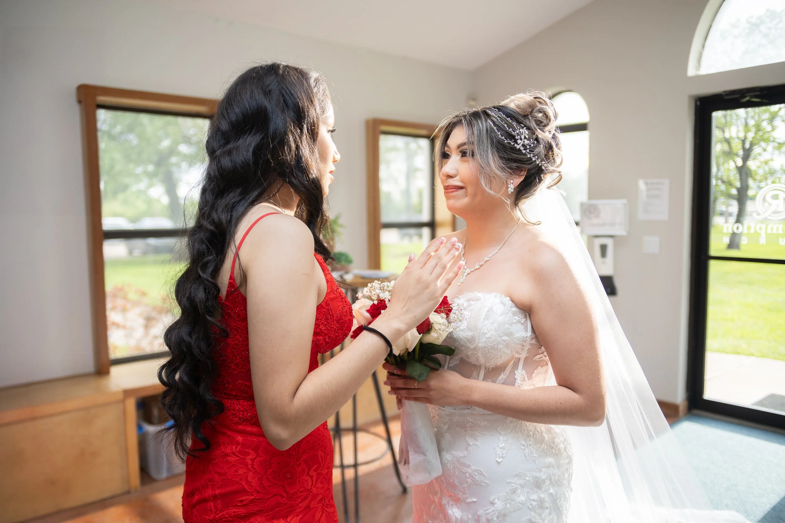 Bride and bridesmaid sharing a heartfelt moment indoors, with the bride holding a bouquet of roses and wearing a white wedding dress, while the bridesmaid wears a red dress.