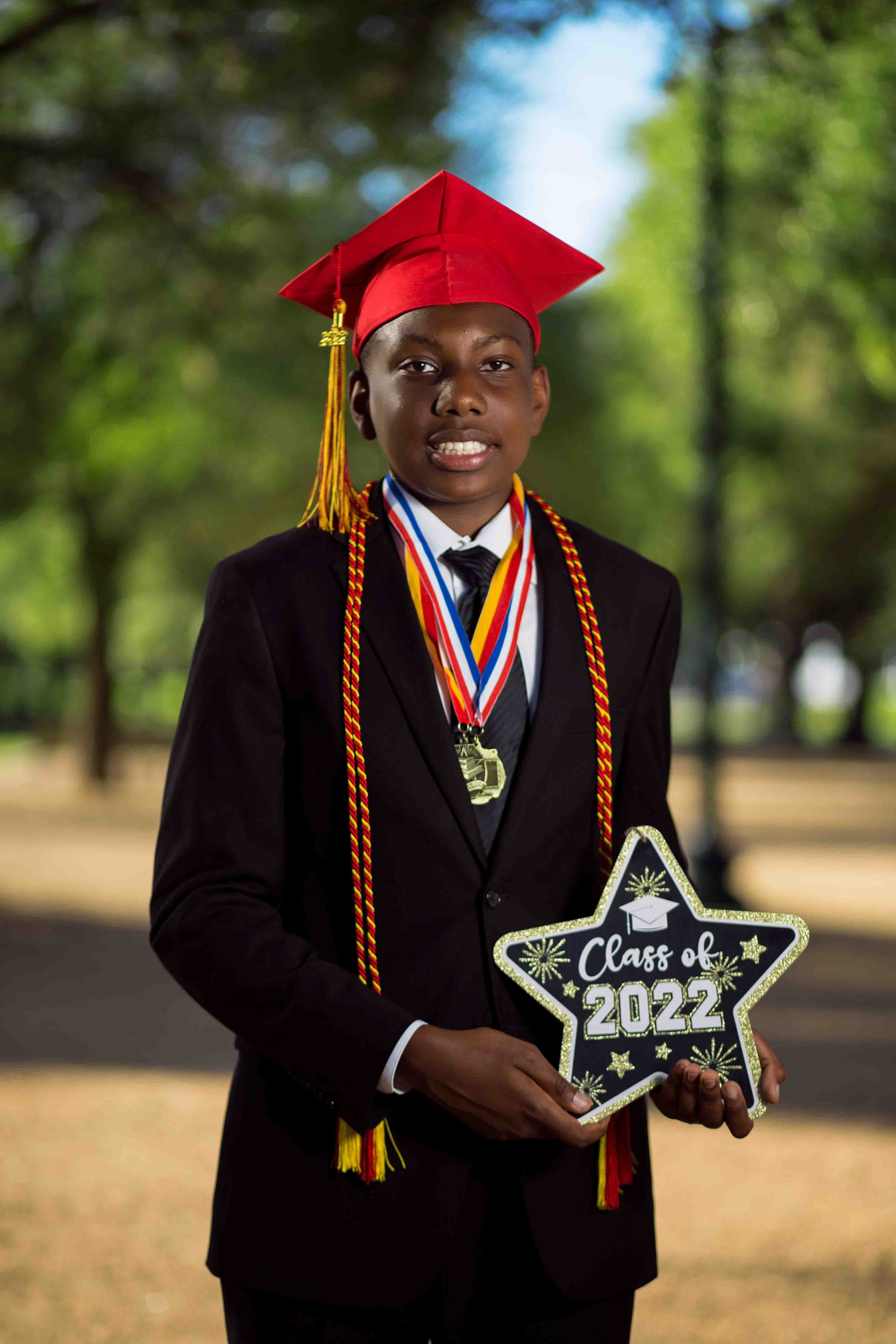 Outdoor graduation portrait of a young boy in a black suit and red graduation cap holding a Christmas-shaped sign that reads “Class of 2022,” wearing medals around his neck in a park setting