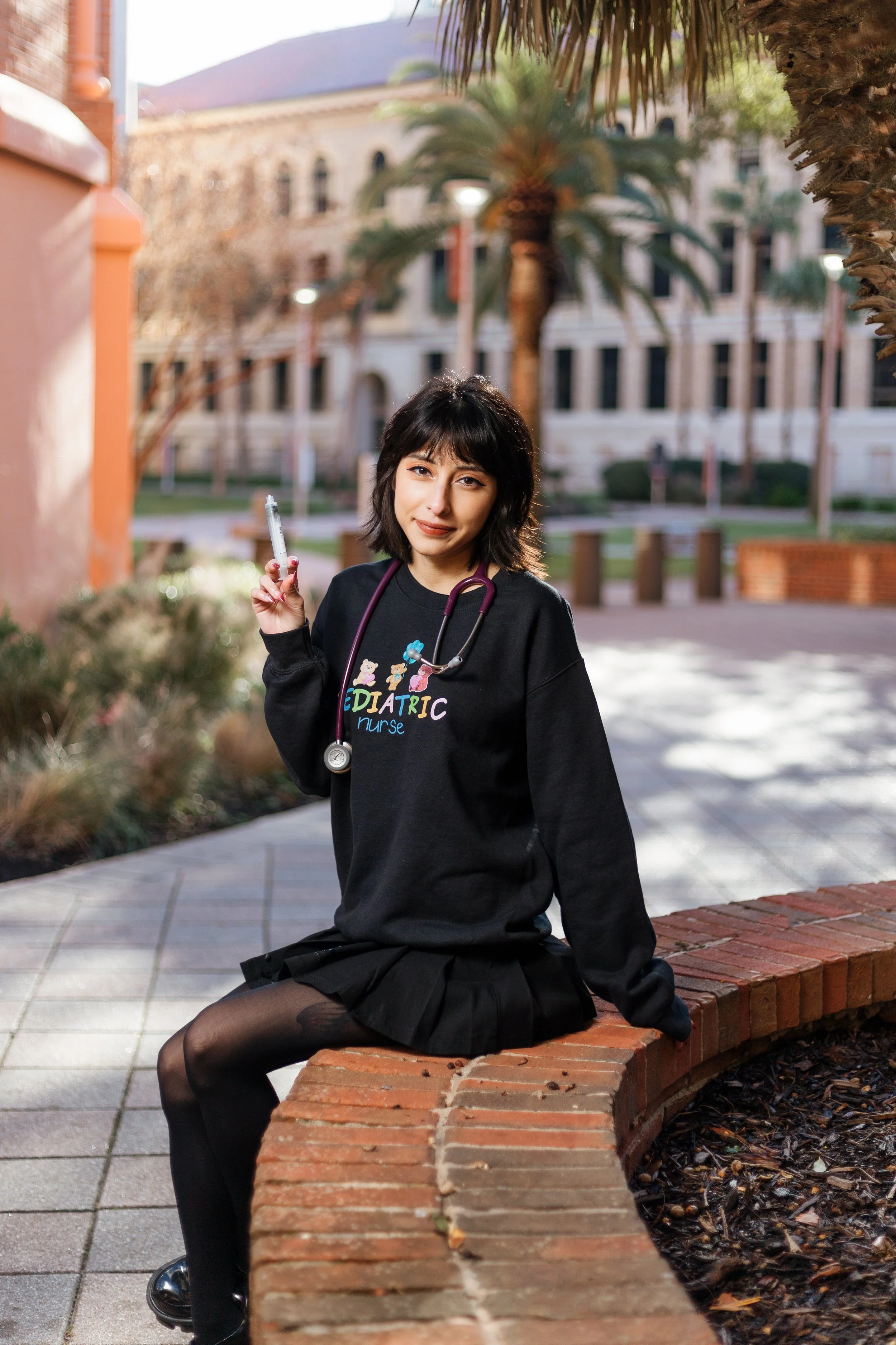 A young woman dressed in a black sweatshirt and skirt, sitting on a brick bench outdoors, holding a syringe, with a stethoscope around her neck, in front of a university building with palm trees.