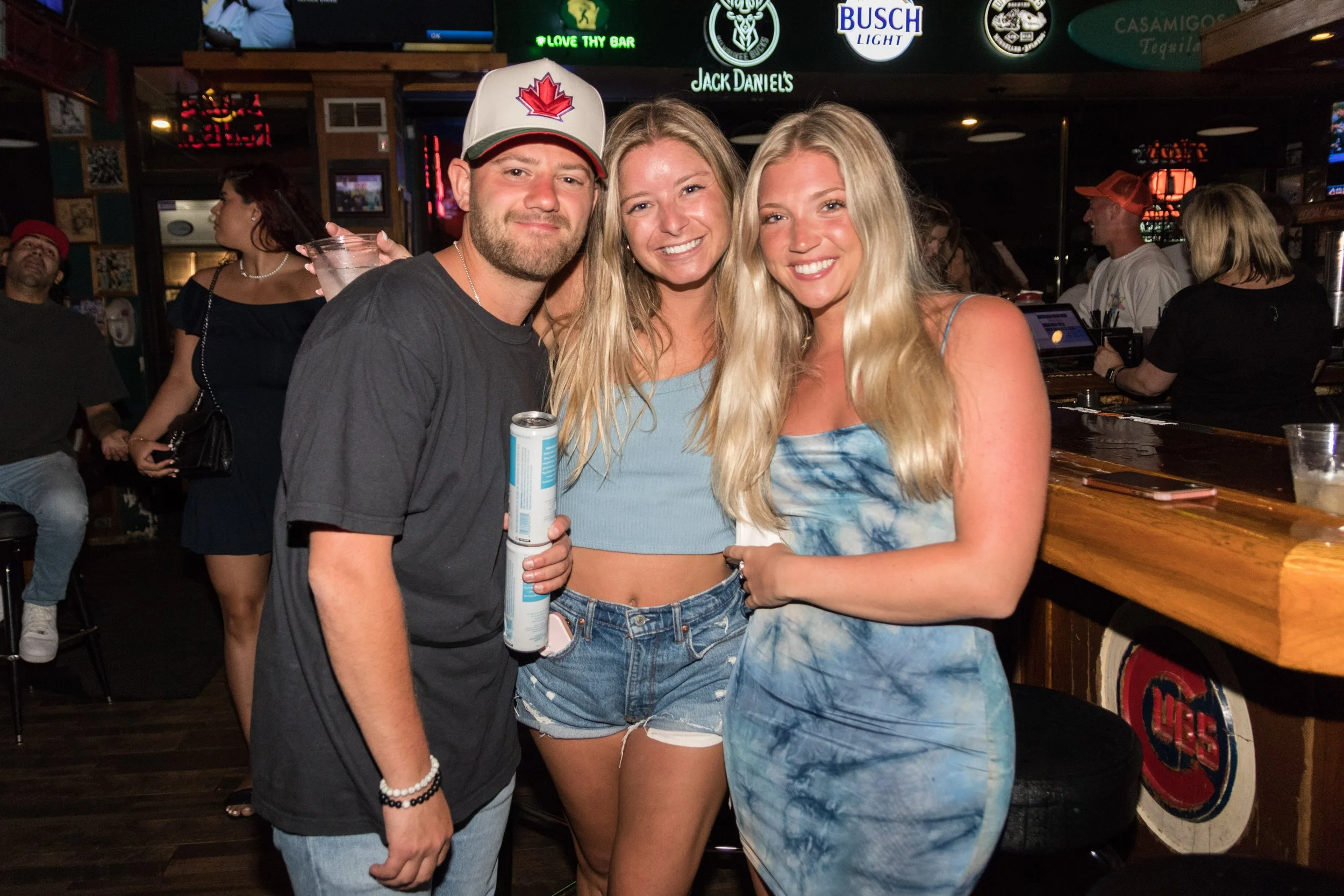 Three young adults, one man and two women, smiling and posing together at a bar. The man is holding a canned drink and wearing a baseball cap with a red maple leaf. The women have long blonde hair, and they are wearing summer tops; one is in a tie-dy