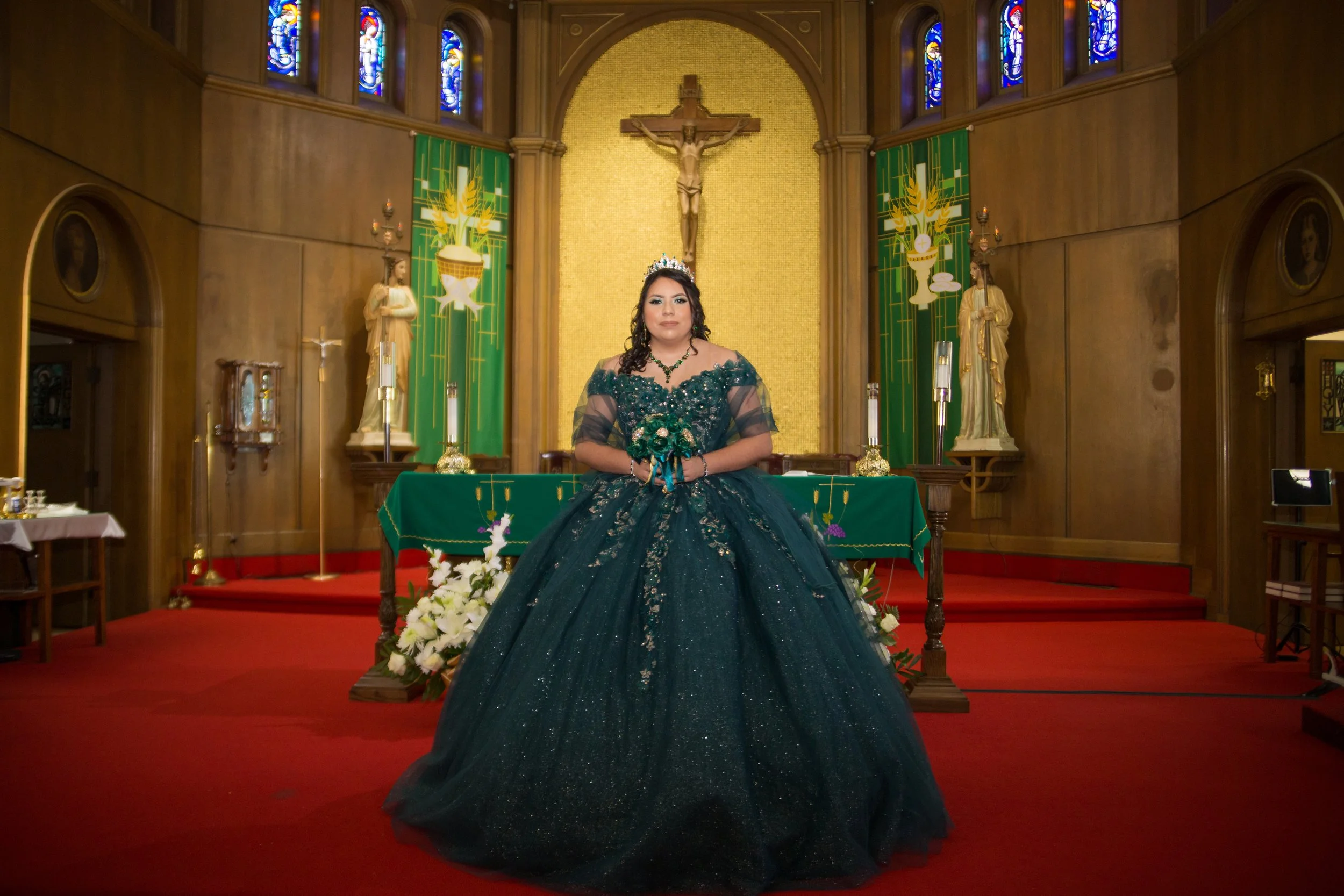 A woman dressed in an elegant, dark green gown standing inside a church with wooden walls, stained glass windows, religious statues, and a crucifix behind her.