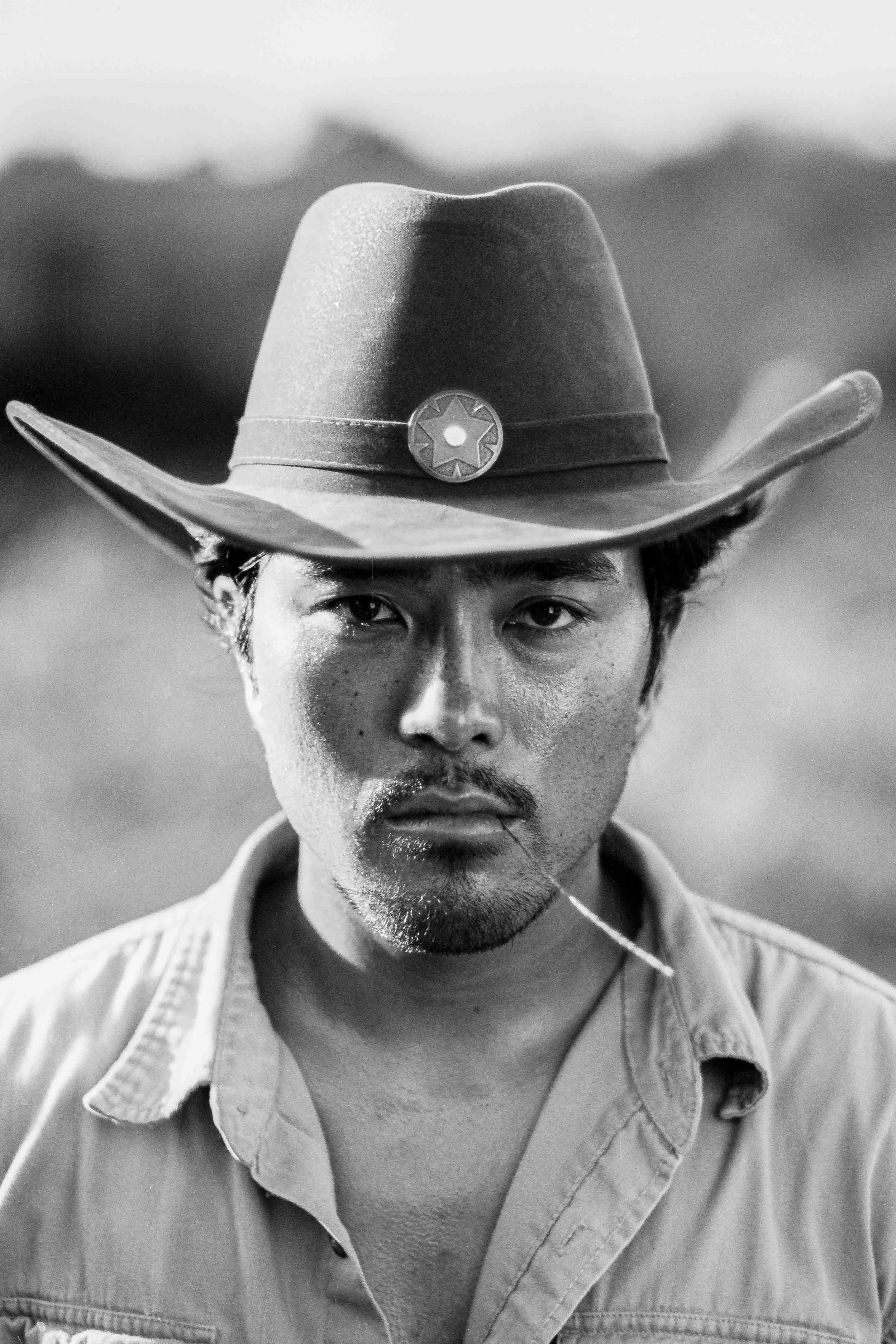 Black and white 35mm film-style portrait of a man wearing a cowboy hat with a badge, looking directly at the camera with a serious expression in a western-inspired setting