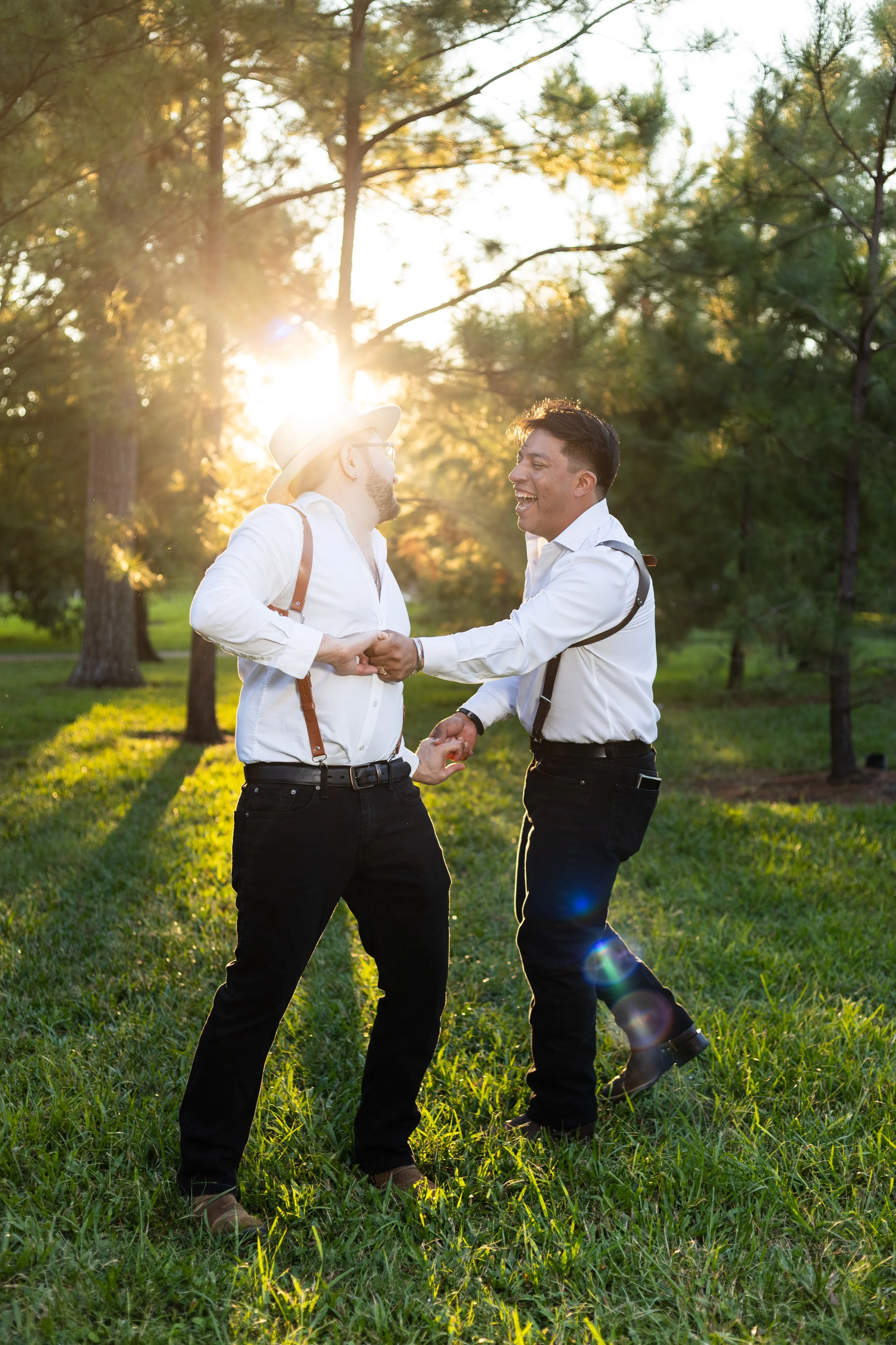 Two men dressed in white shirts and black pants, holding hands and laughing in a grassy park with trees and sunlight in the background.