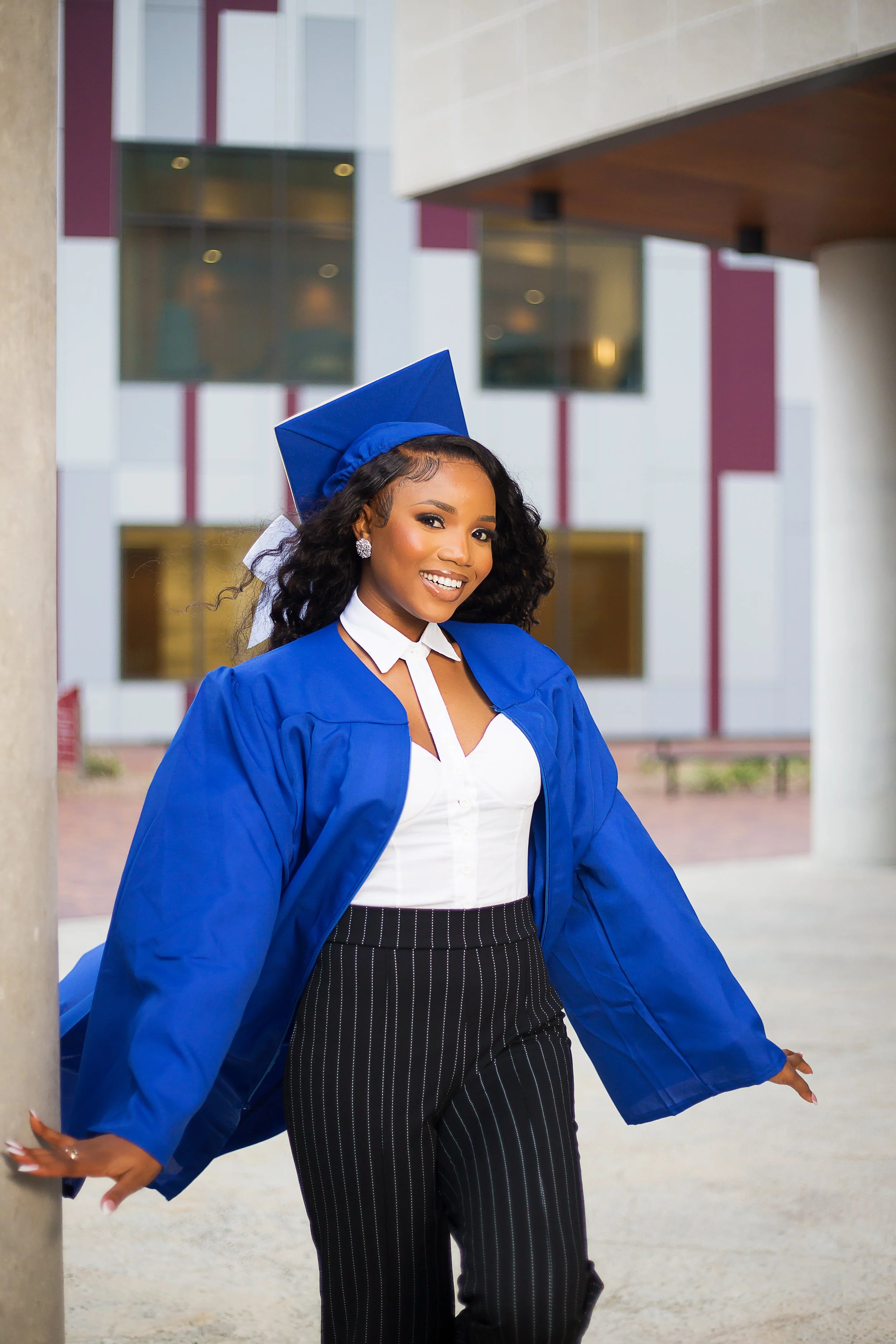 Young woman in a blue graduation gown and cap, smiling, standing outdoors in front of a modern building.