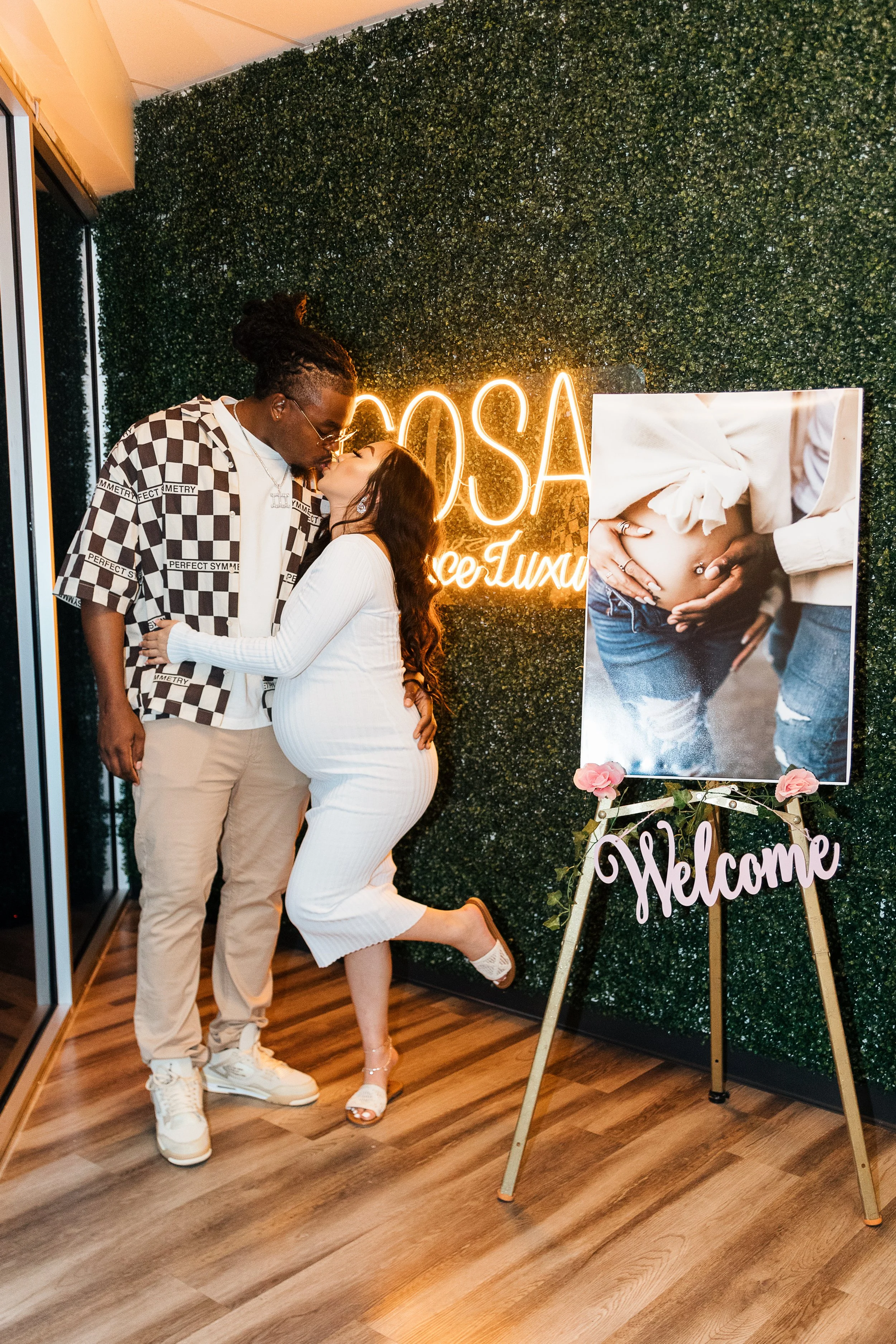 Couple kissing at a baby shower, with a neon sign saying 'Baby Shower', a poster of a pregnant woman, and a 'Welcome' sign with pink flowers.