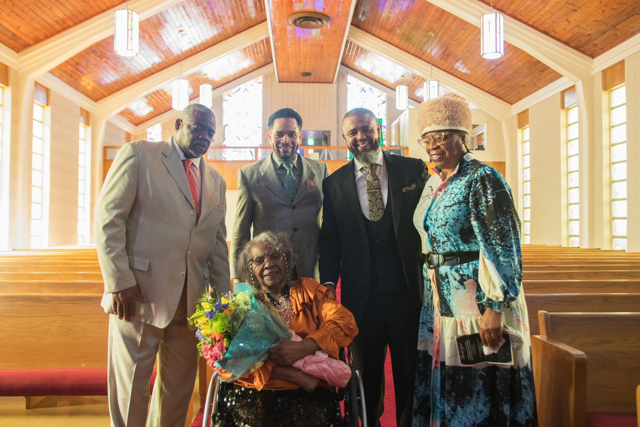 Group of six people, five standing and one seated in a wheelchair holding a bouquet of flowers, inside a church with wooden pews and stained glass windows.