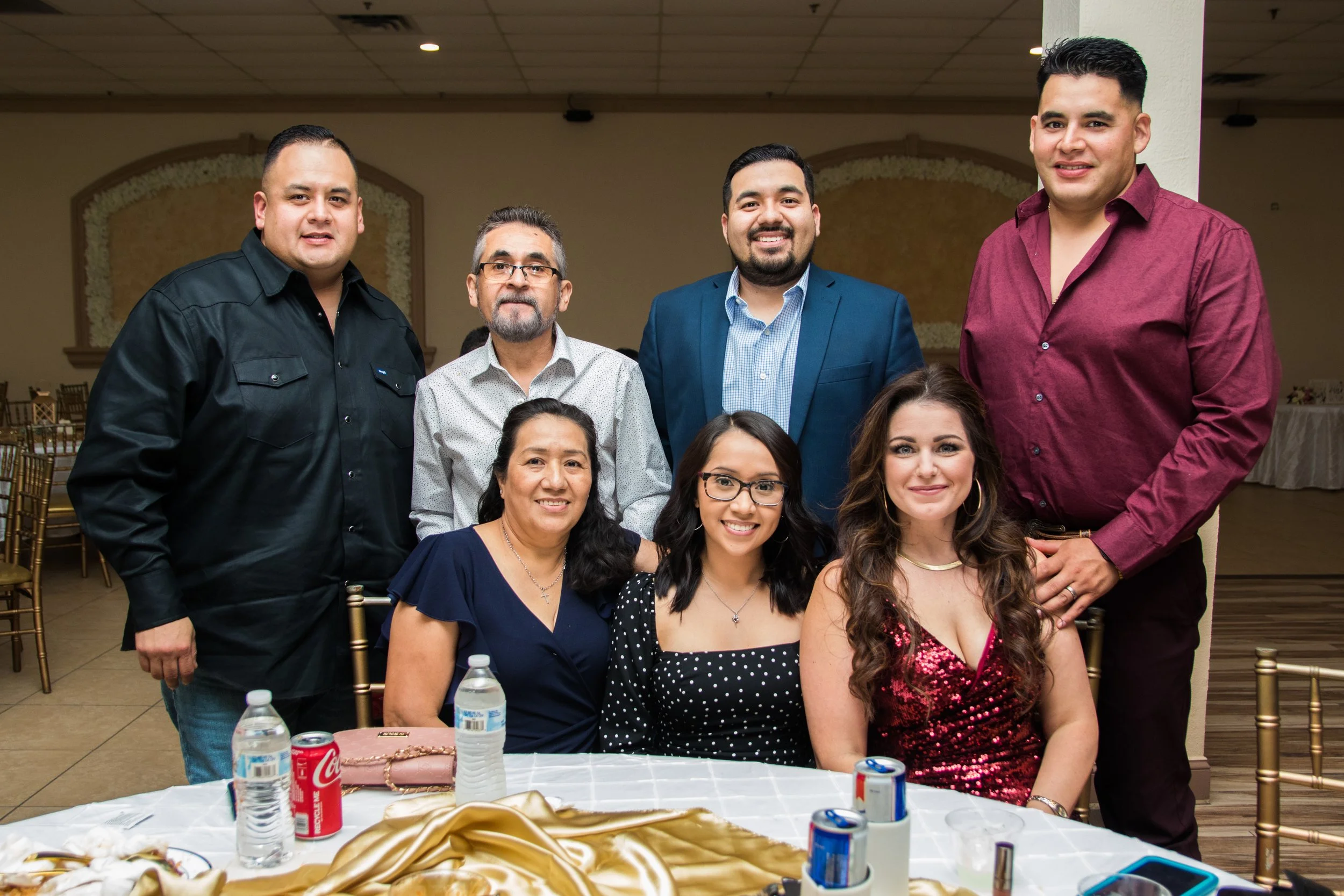 Group of seven diverse people smiling at a formal event, seated and standing around a decorated table with drinks and food.