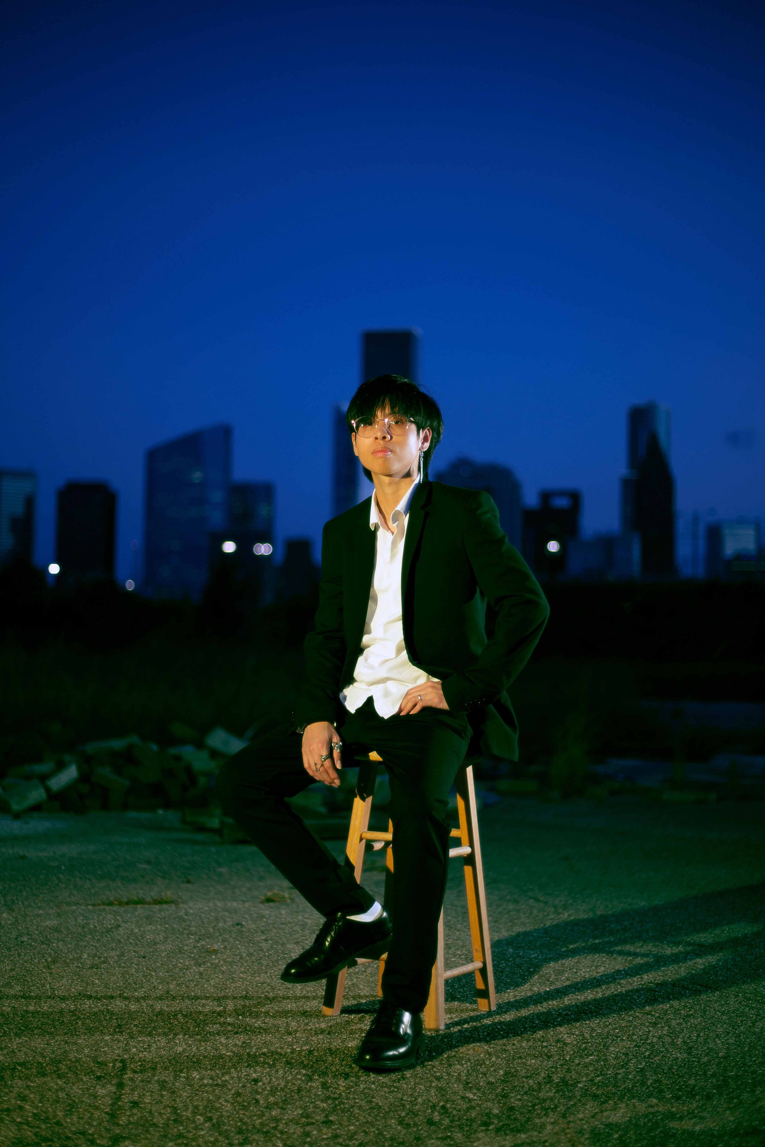 Downtown Houston nighttime portrait by Lev’s Photography featuring a person in a suit sitting on a stool outdoors with the city skyline in the background for an album cover session