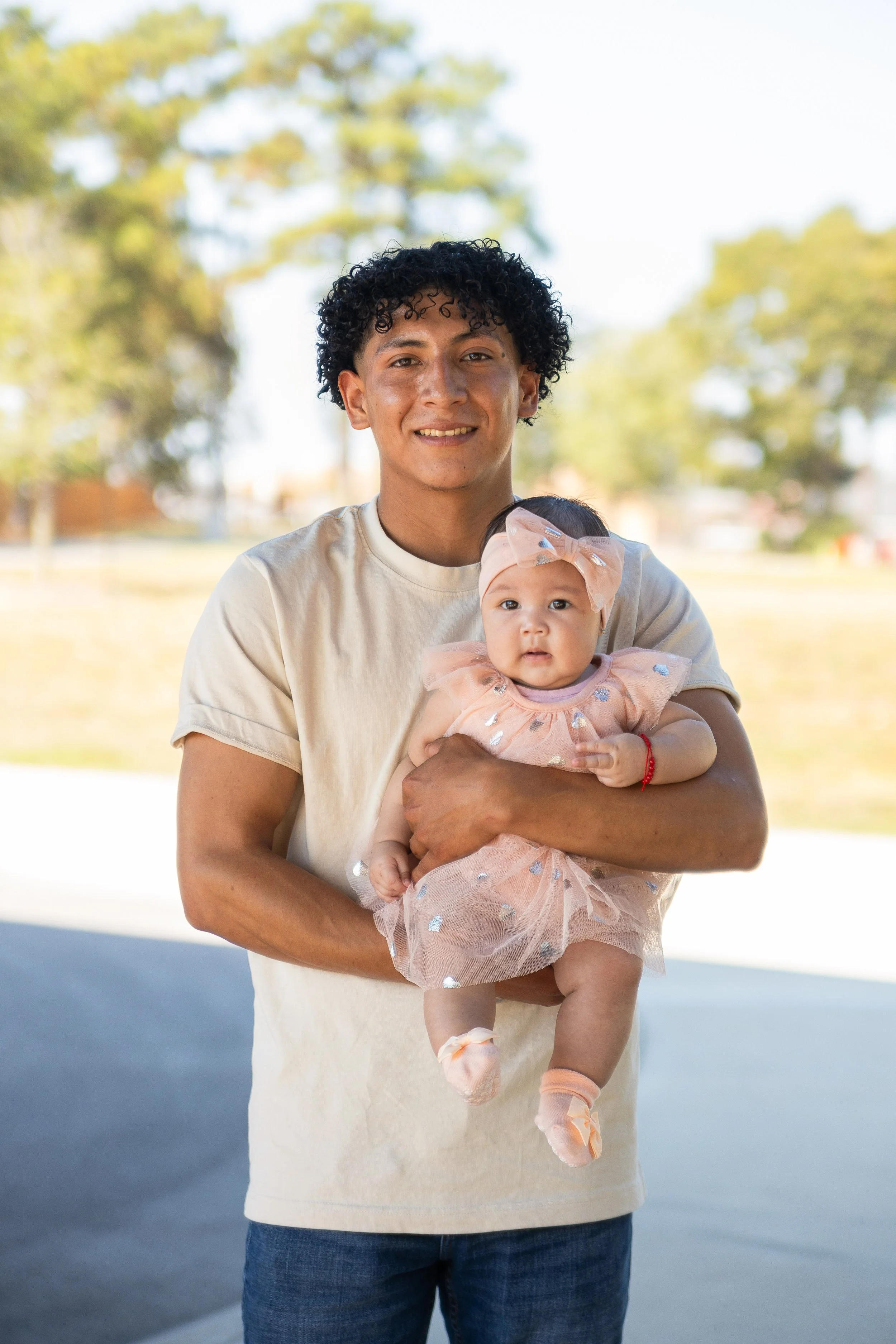 A man holding a young girl wearing a pink dress with shiny embellishments and a large pink bow headband, outdoors on a sunny day with trees in the background.