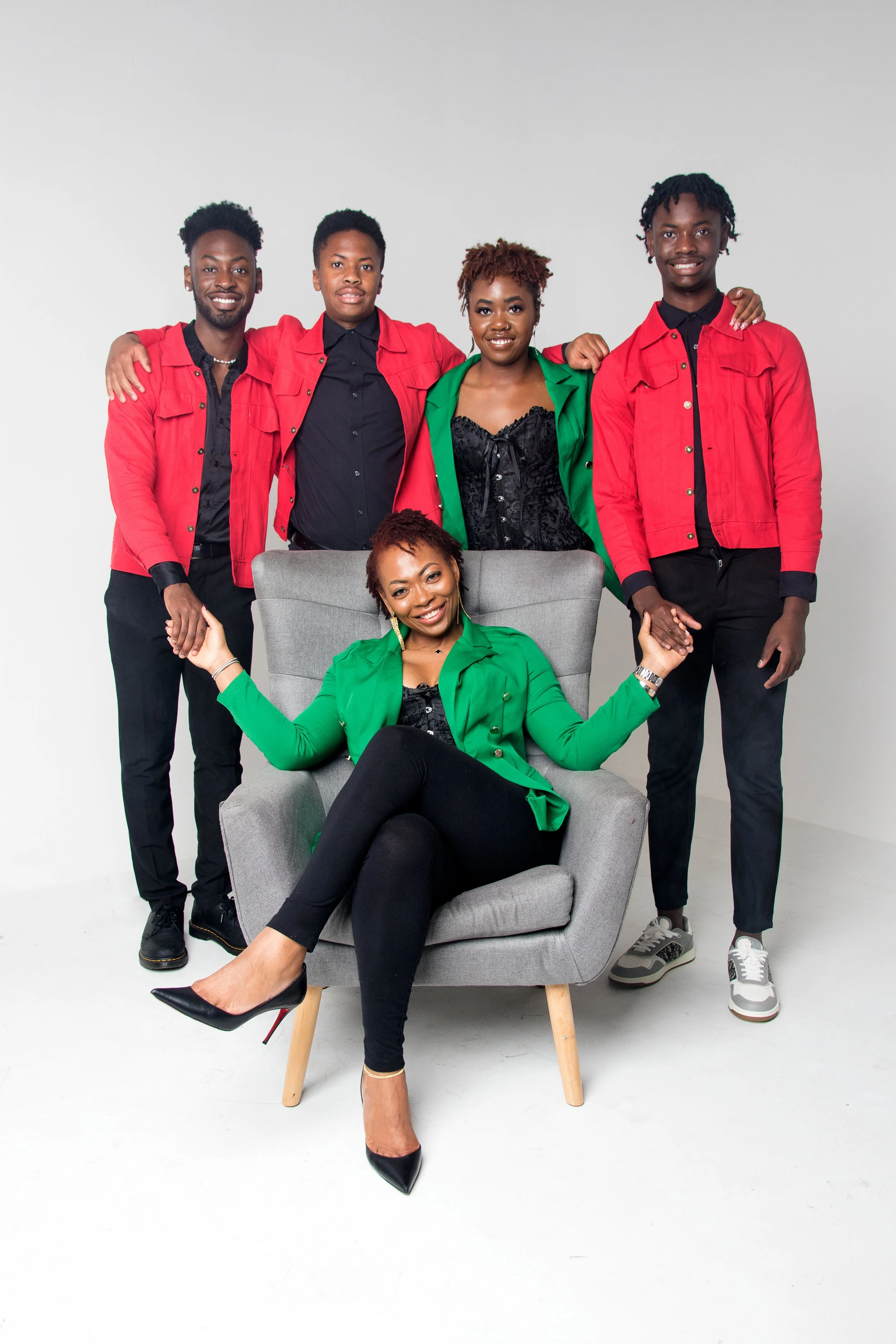 Group of five diverse young adults, four men and one woman, posing together with the woman seated in a gray armchair. Three of the men stand behind her, and she is smiling with arms raised, wearing a green blazer and black pants, in a studio with pla