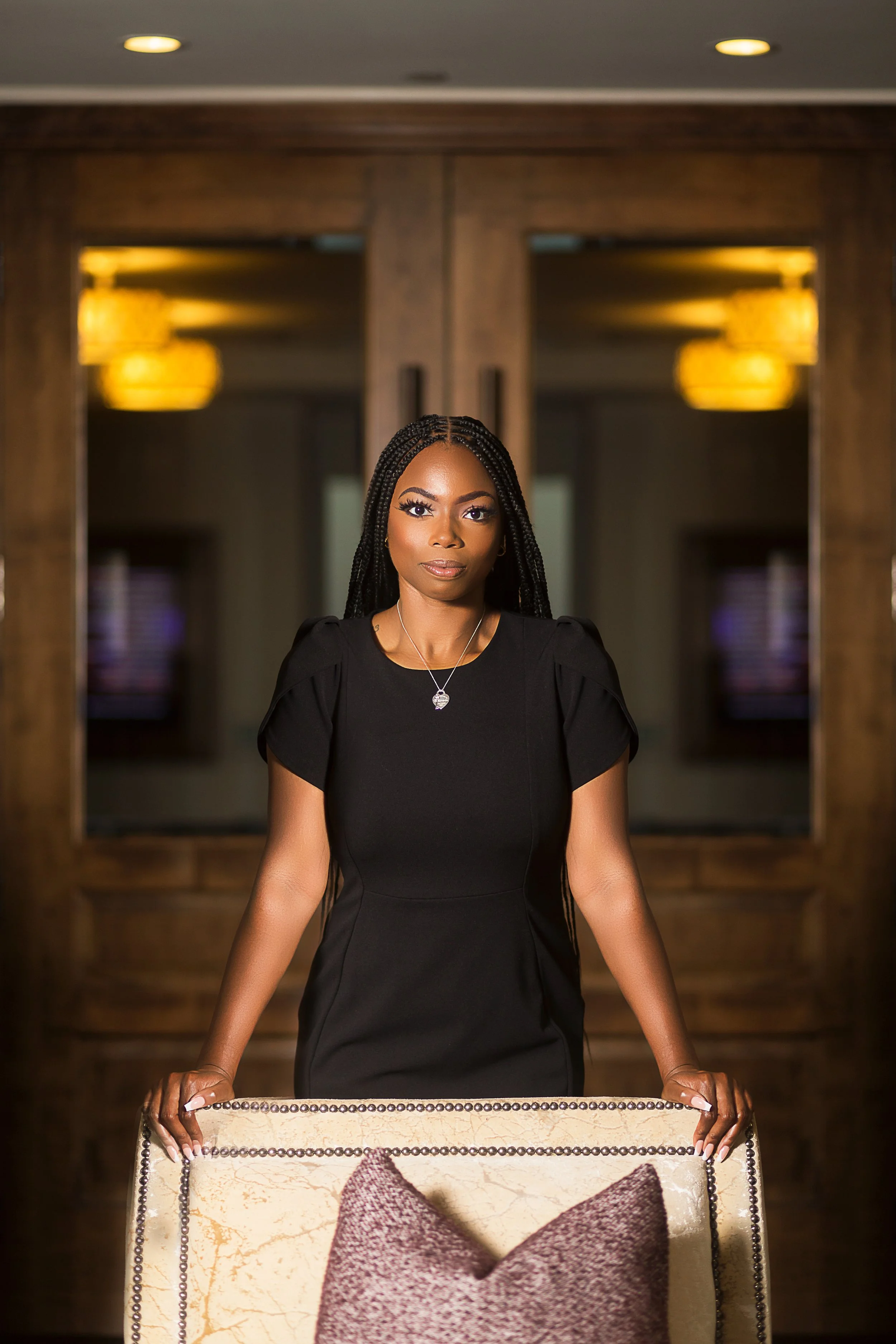 A woman with braided hair wearing a black dress, standing behind a chair in an indoor setting with wooden doors and warm lighting.