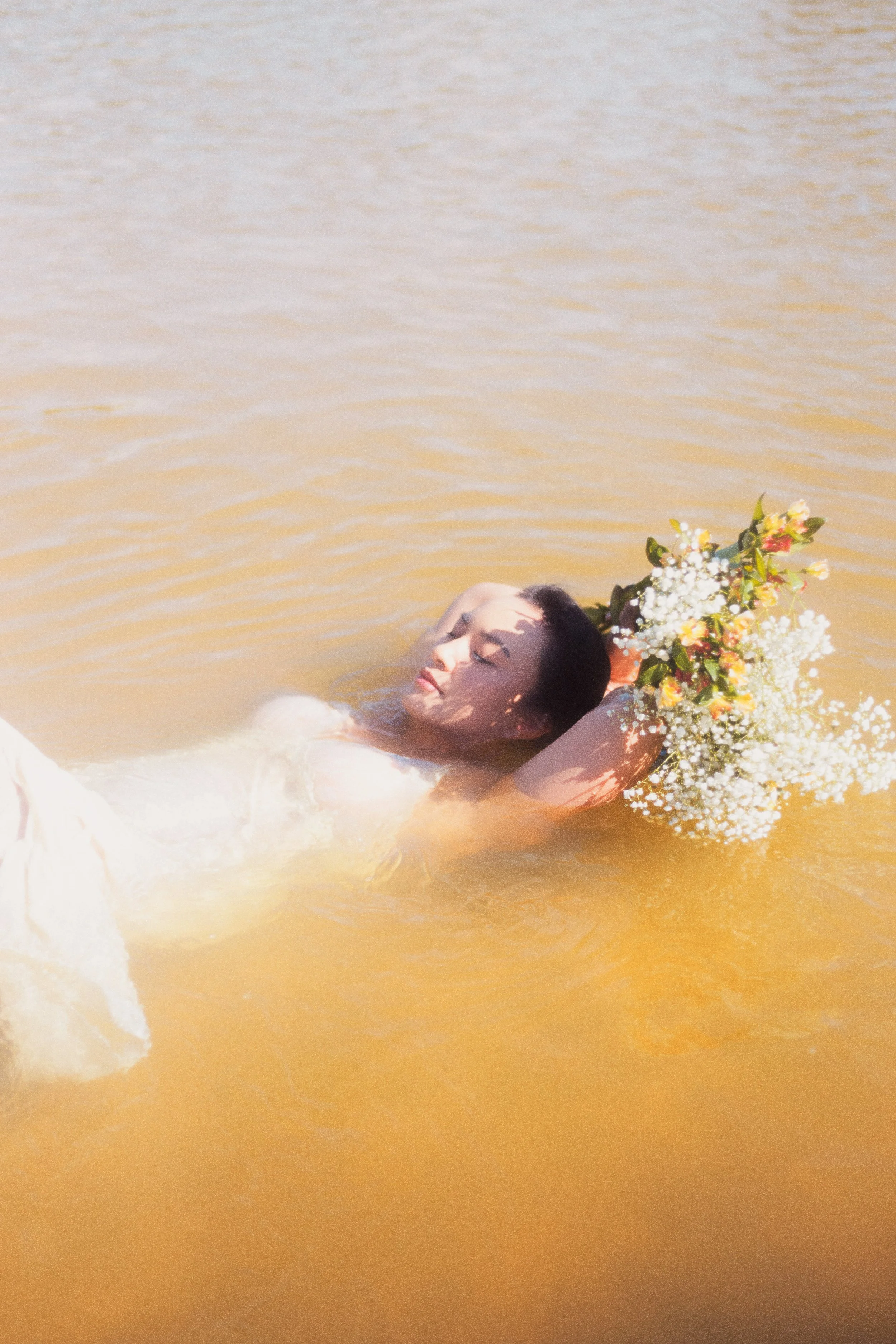 A woman floats in water with eyes closed, holding a bouquet of flowers behind her head.