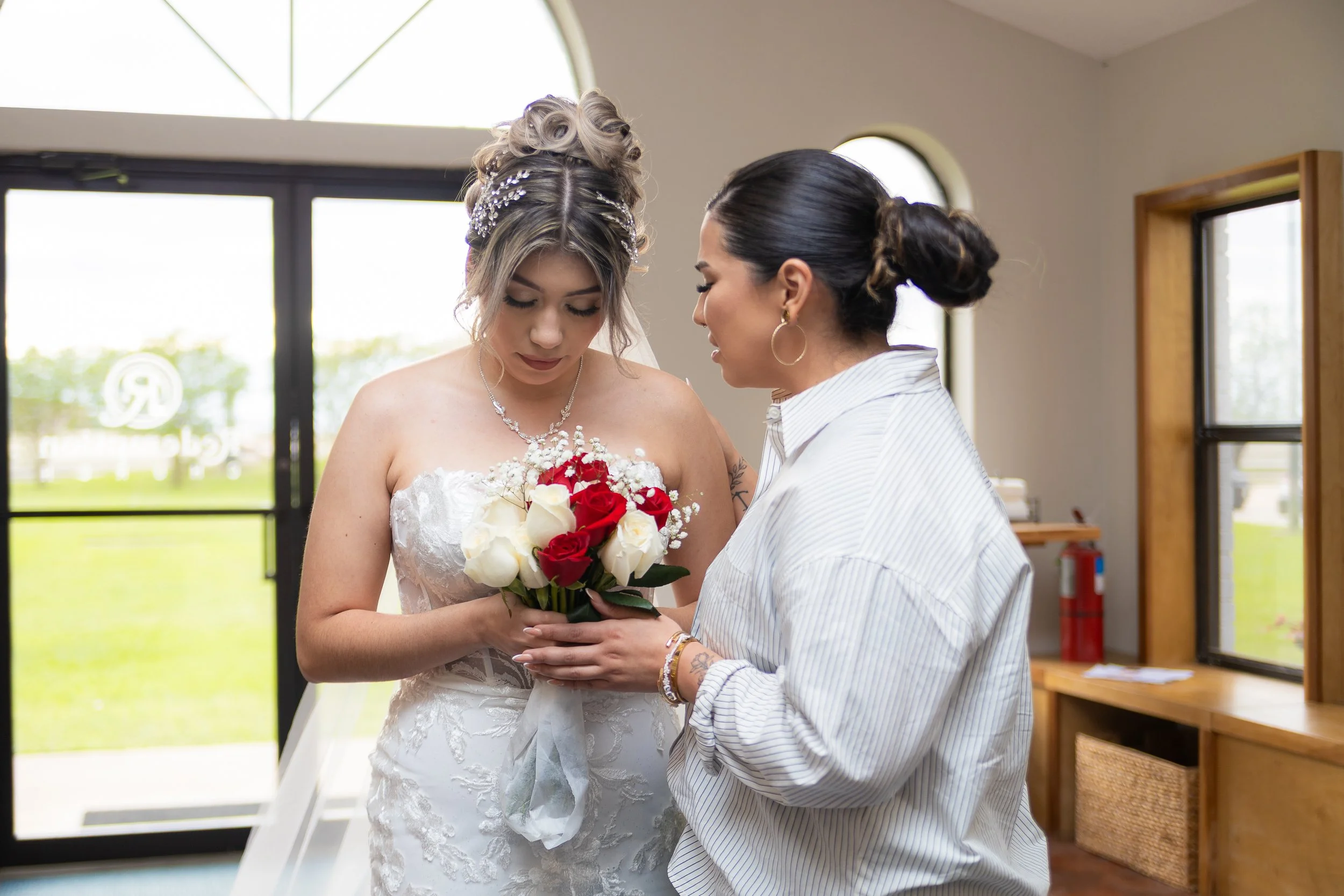 A bride in a wedding dress holding a bouquet of red and white roses, standing indoors near a window, while a woman dressed in a striped shirt appears to be adjusting or giving her something.