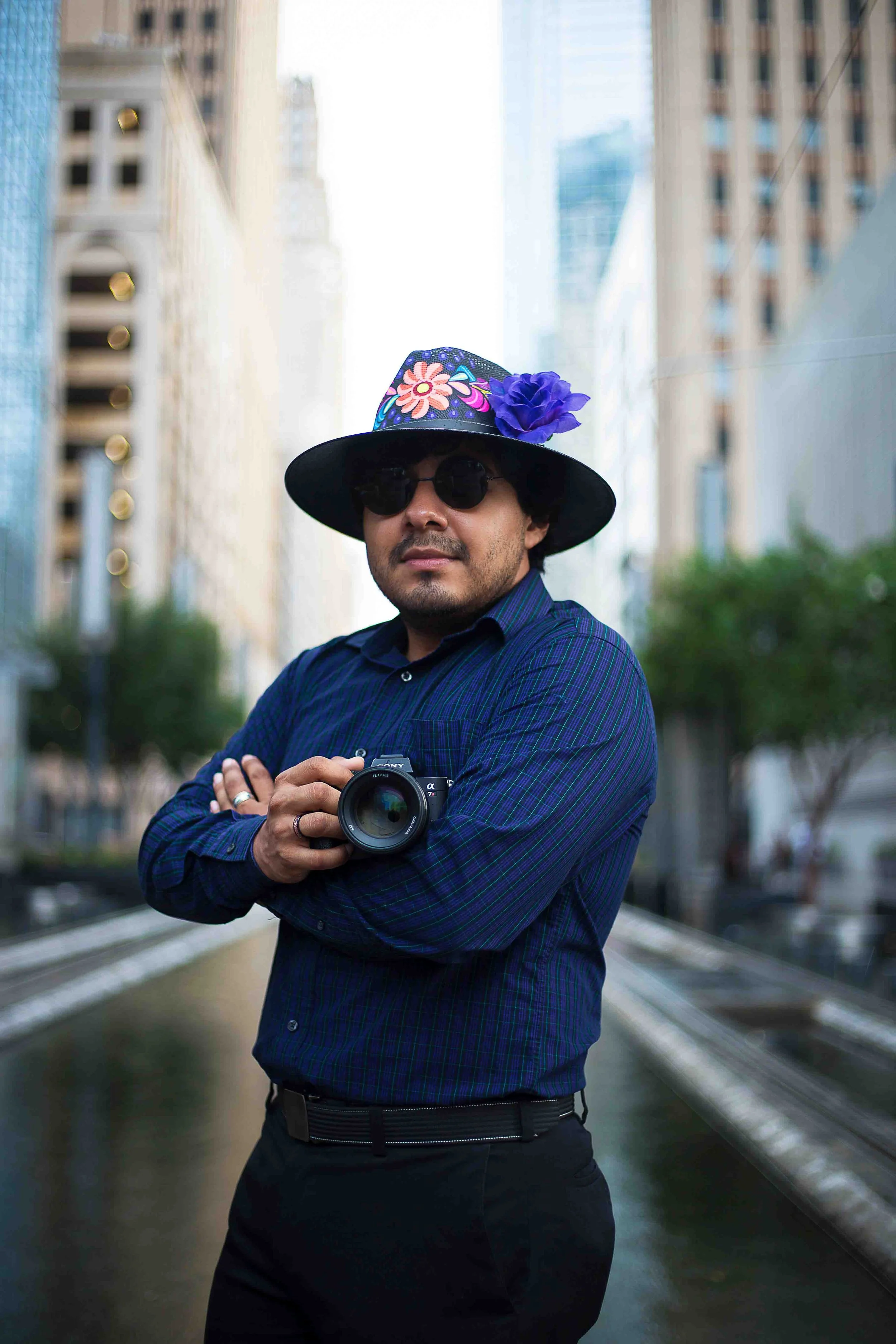 Downtown Houston editorial portrait by Lev’s Photography featuring a man in a floral hat, sunglasses, and dark blue plaid shirt holding a camera outdoors with tall buildings and trees in the background for a newspaper photoshoot