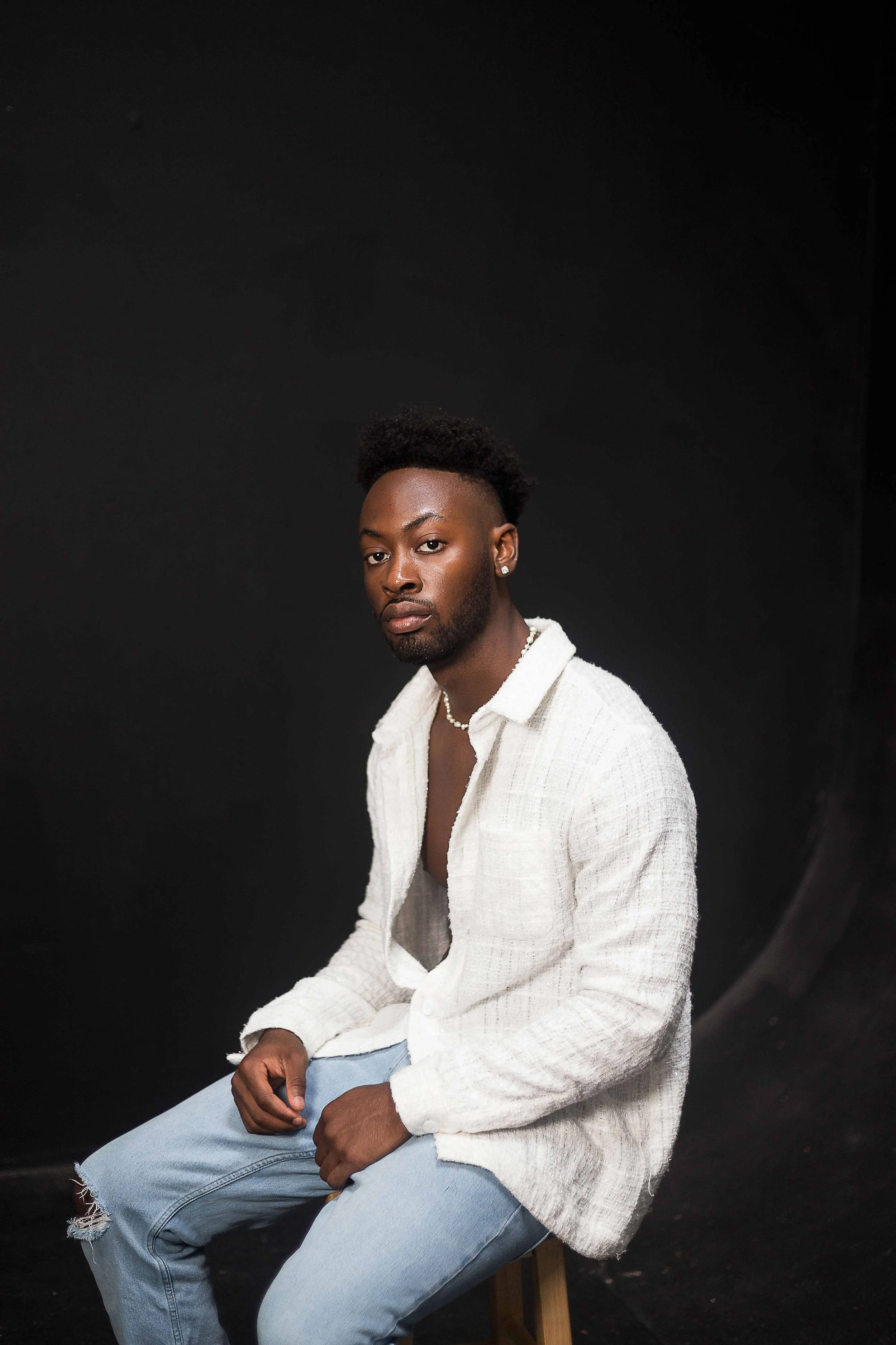 Studio birthday portrait in Houston, Texas by Lev’s Photography featuring a young man with dark skin and short curly hair wearing a white textured shirt, ripped light blue jeans, and pearl accessories against a black background