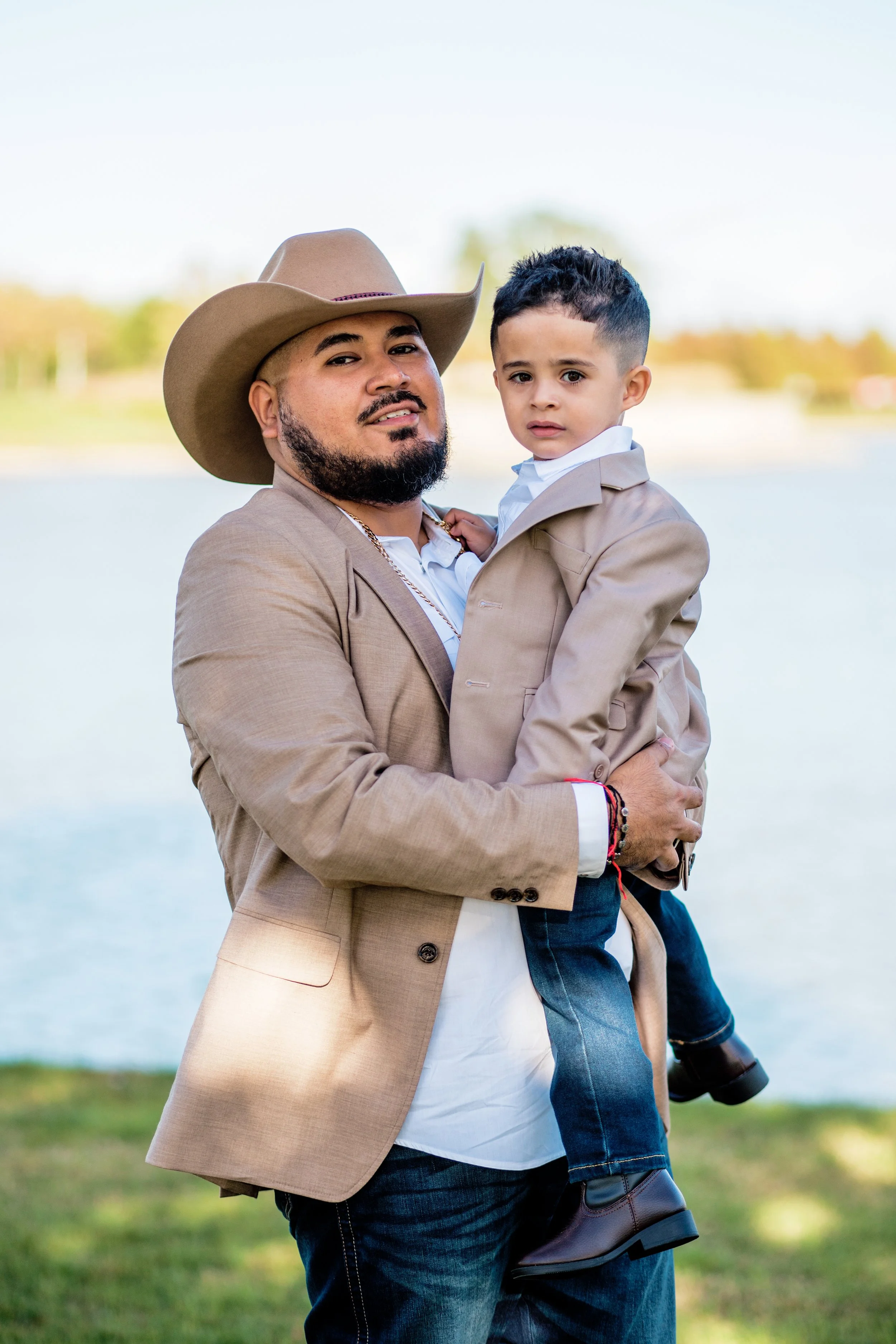 A man in a tan blazer and cowboy hat holding a young boy in similar attire near a body of water with trees in the background.