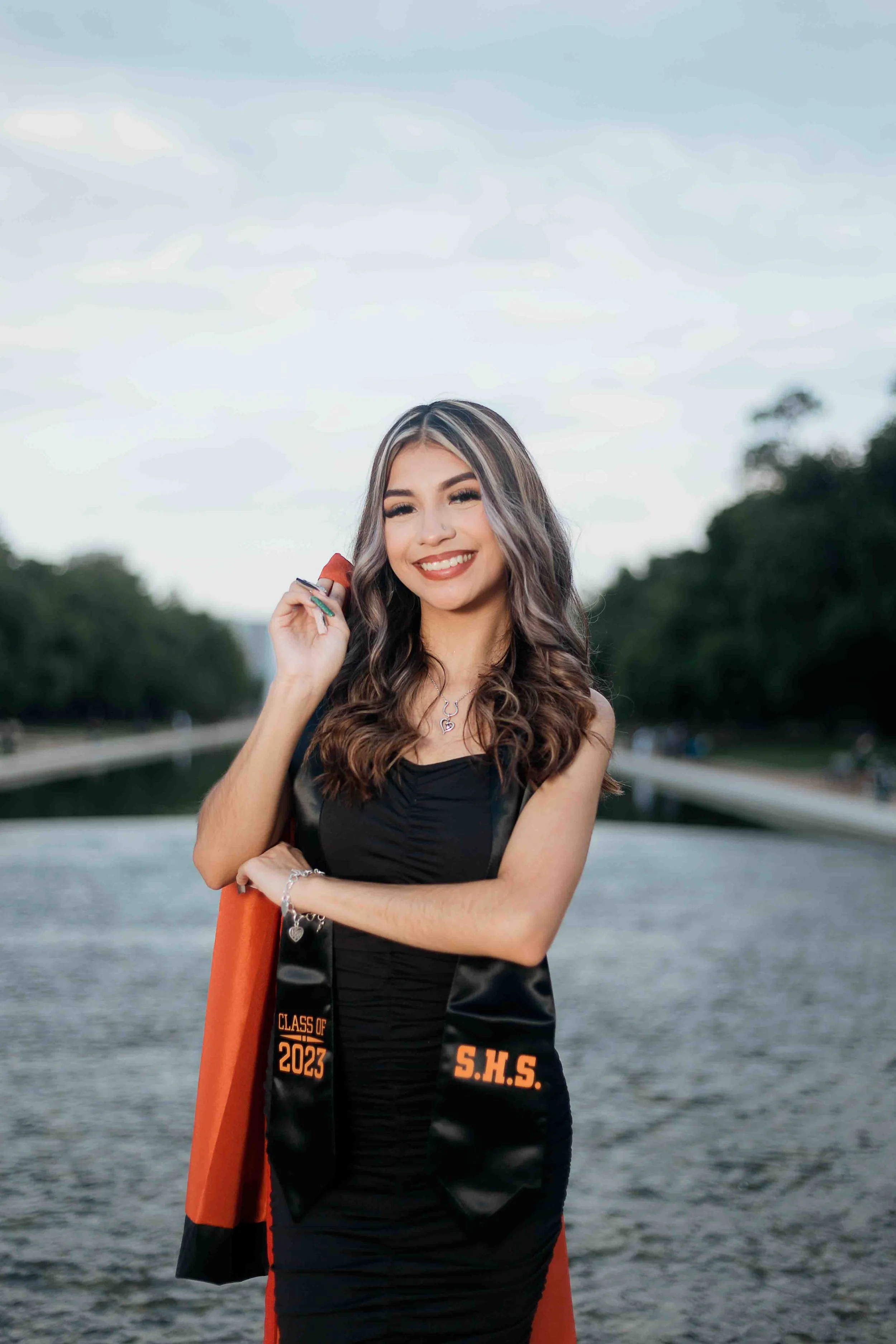 Graduation portrait of a young woman in a black dress and a black-and-orange stole standing outdoors by a waterway, smiling and holding her graduation cap, with the stole reading “Class of 2023” and “S.H.S