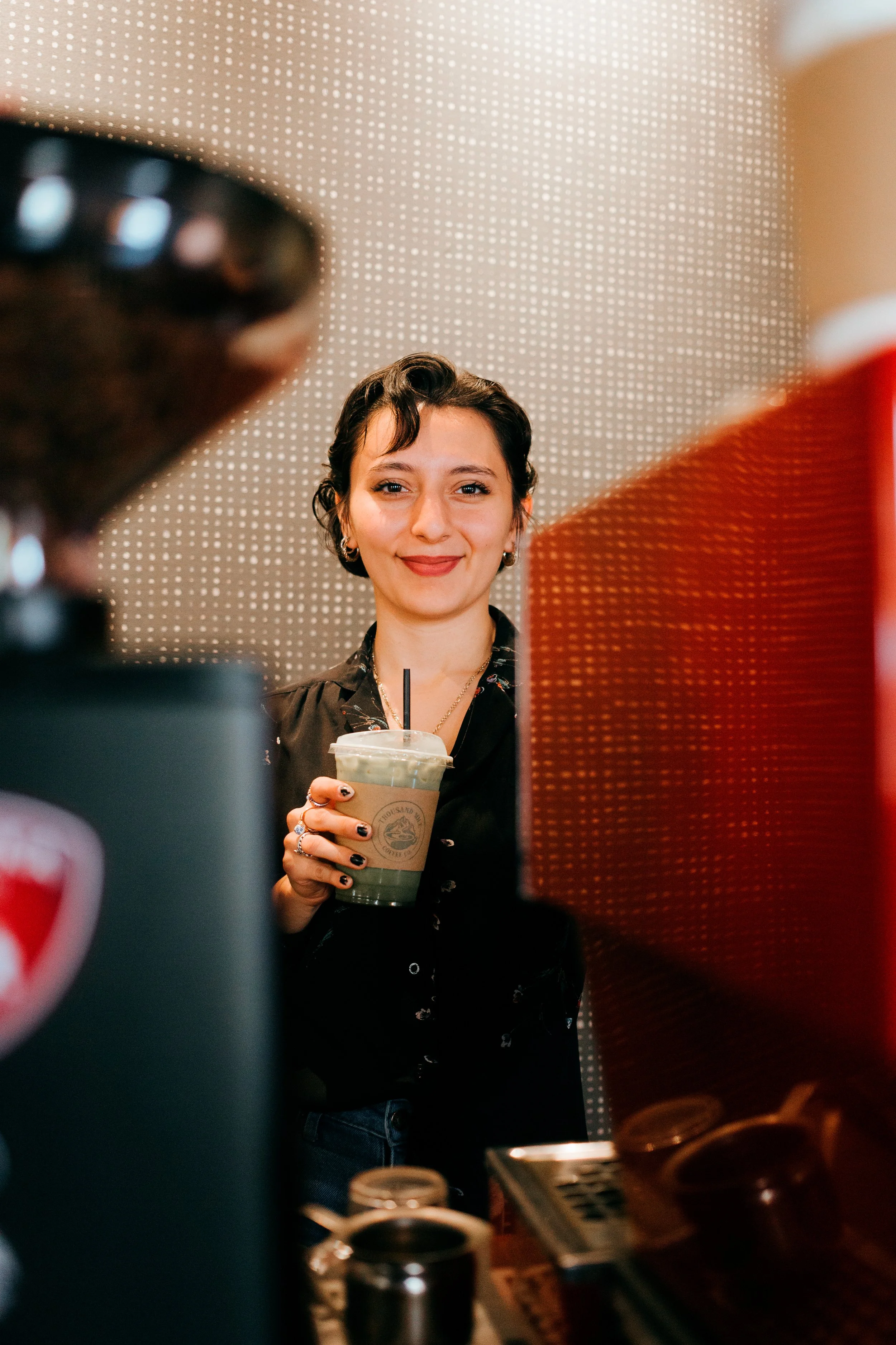 A smiling young woman with short dark hair, holding a plastic cup with a green beverage and a straw, seen through a cafe or coffee shop counter with various equipment and supplies.