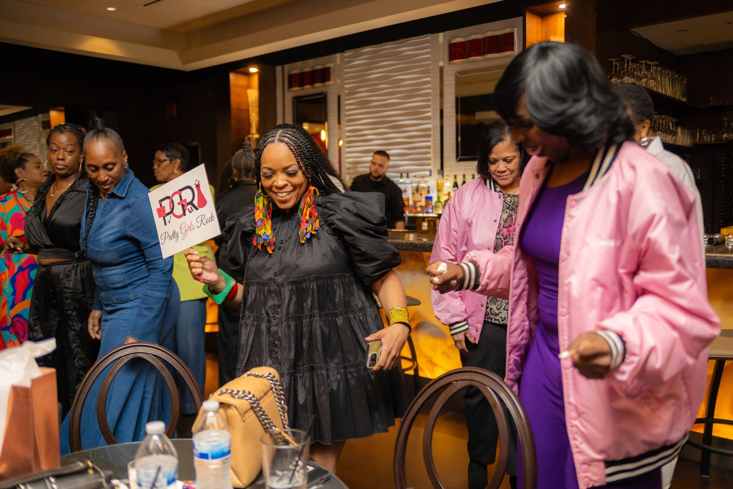 A group of women at an indoor social gathering smiling and interacting, with a bar in the background, some holding drinks and a woman holding a sign that says 'Pretty Girls Rock'.