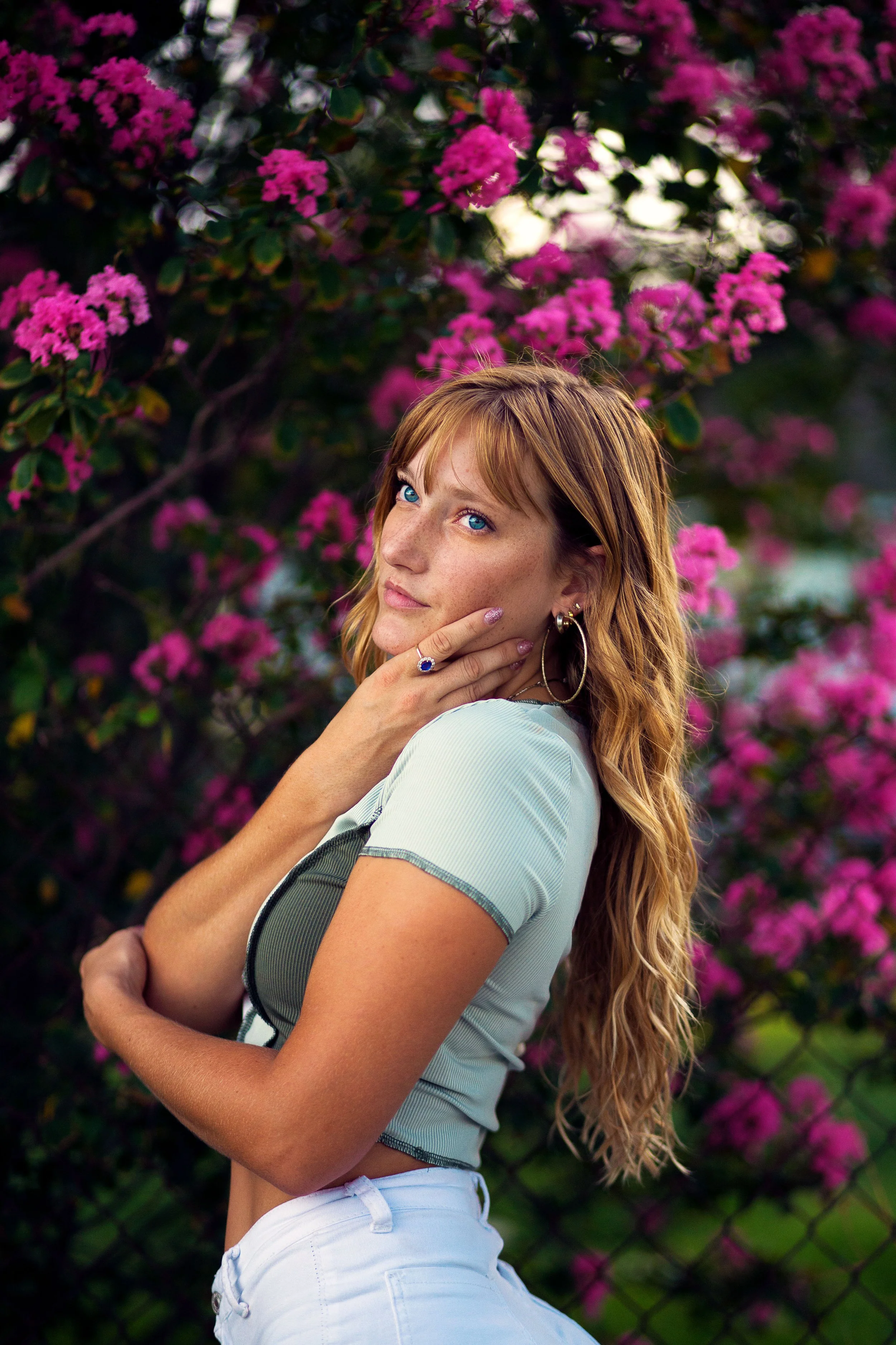 A young woman with long wavy red hair, blue eyes, and freckles, posing outdoors near a flowering bush with pink blossoms. She is wearing earrings, a ring, a light-colored top, and white pants, touching her chin with her right hand and looking into th