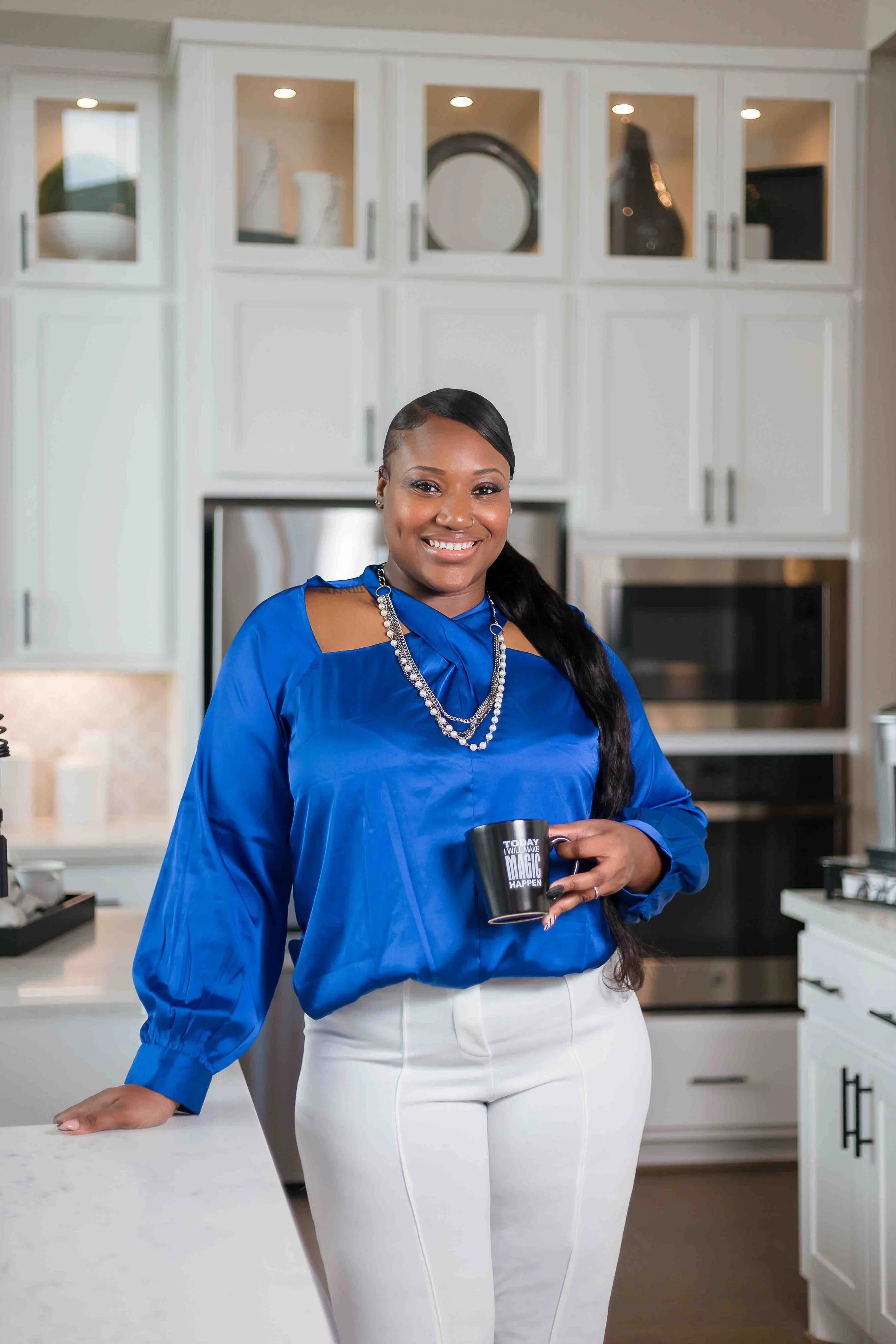 Real estate branding photography in Houston, Texas by Lev’s Photography featuring a smiling realtor in a bright blue blouse holding a “Today I Will Make Magic Happen” mug in a modern kitchen setting