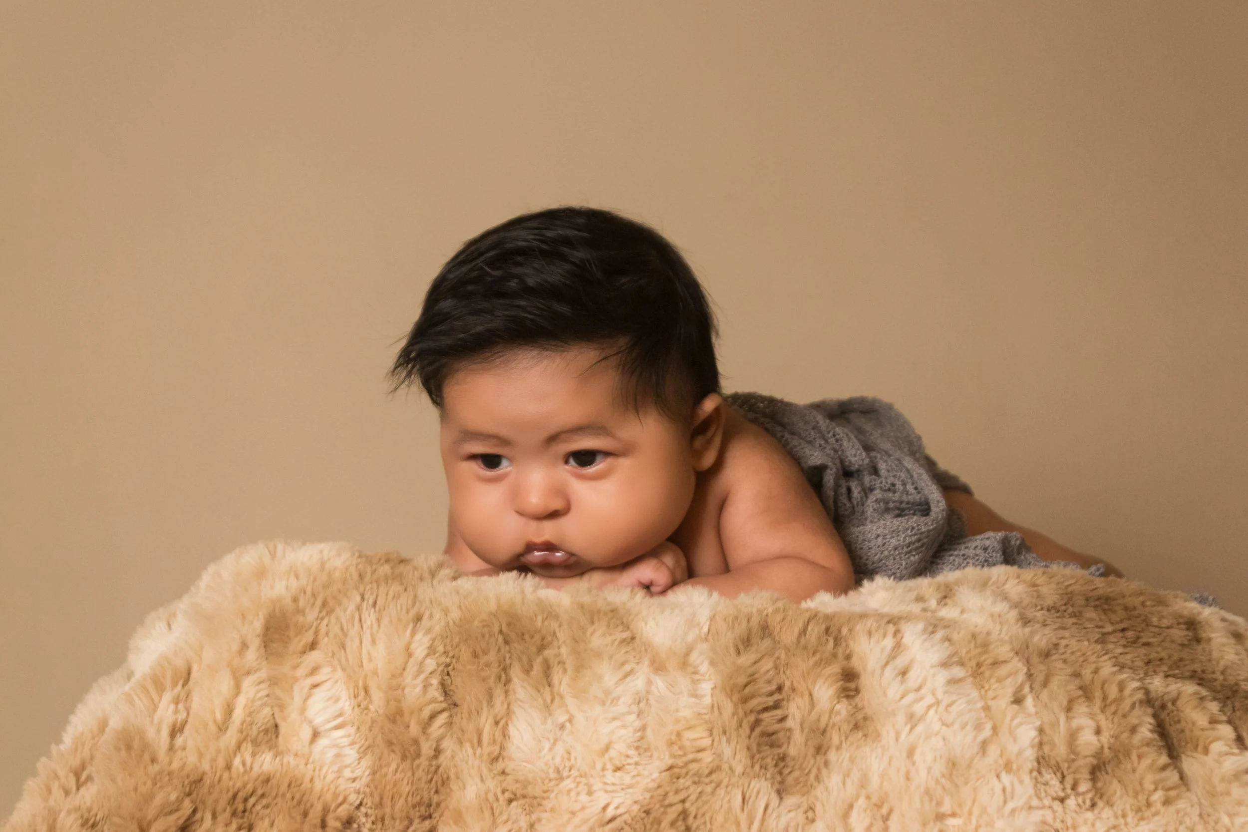 A young child with dark hair lying on stomach on a plush beige blanket, looking intently off camera with a thoughtful expression.