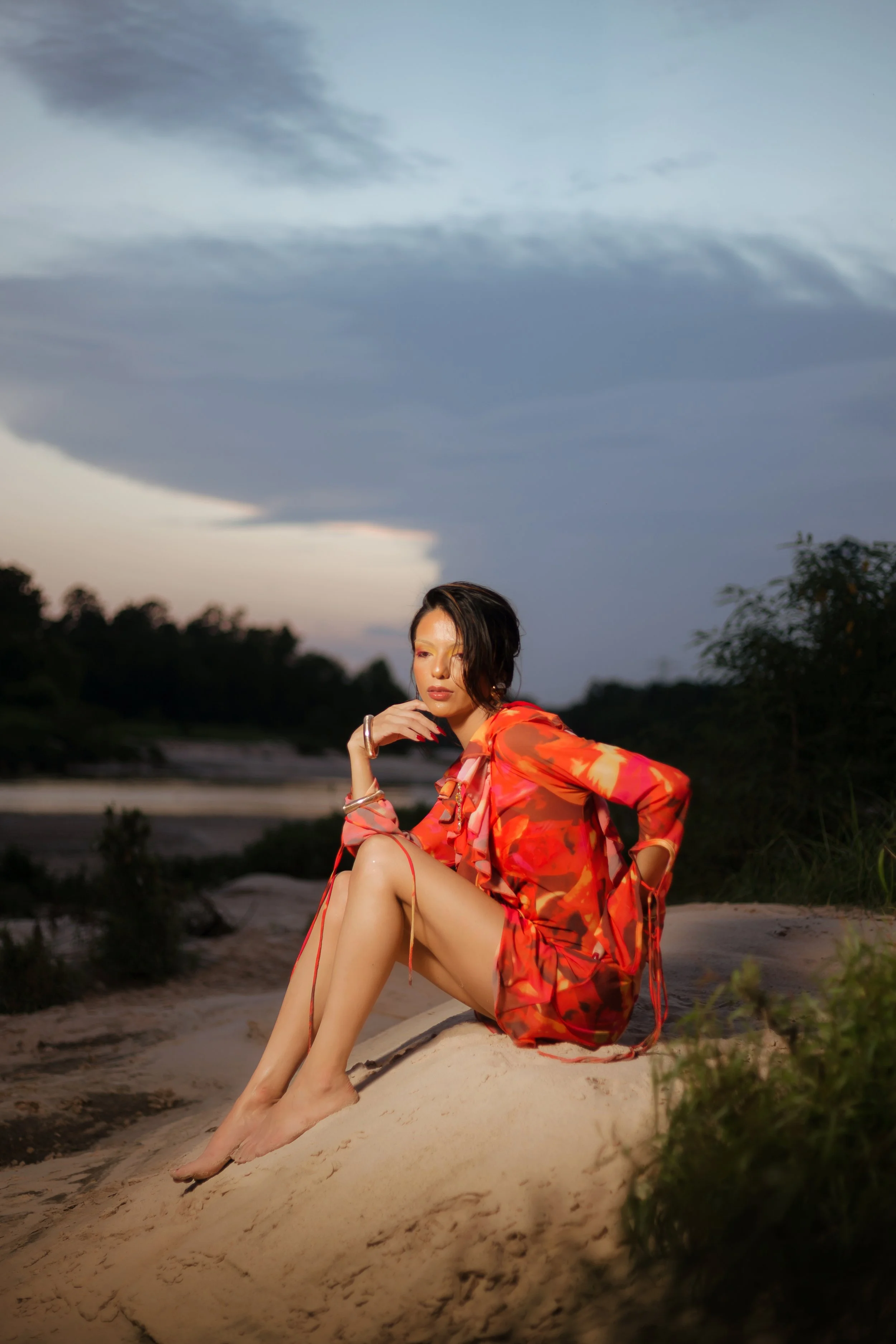 A woman in a vibrant red and orange dress sits on a sandy terrain outdoors during dusk, with a cloudy sky and trees in the background.