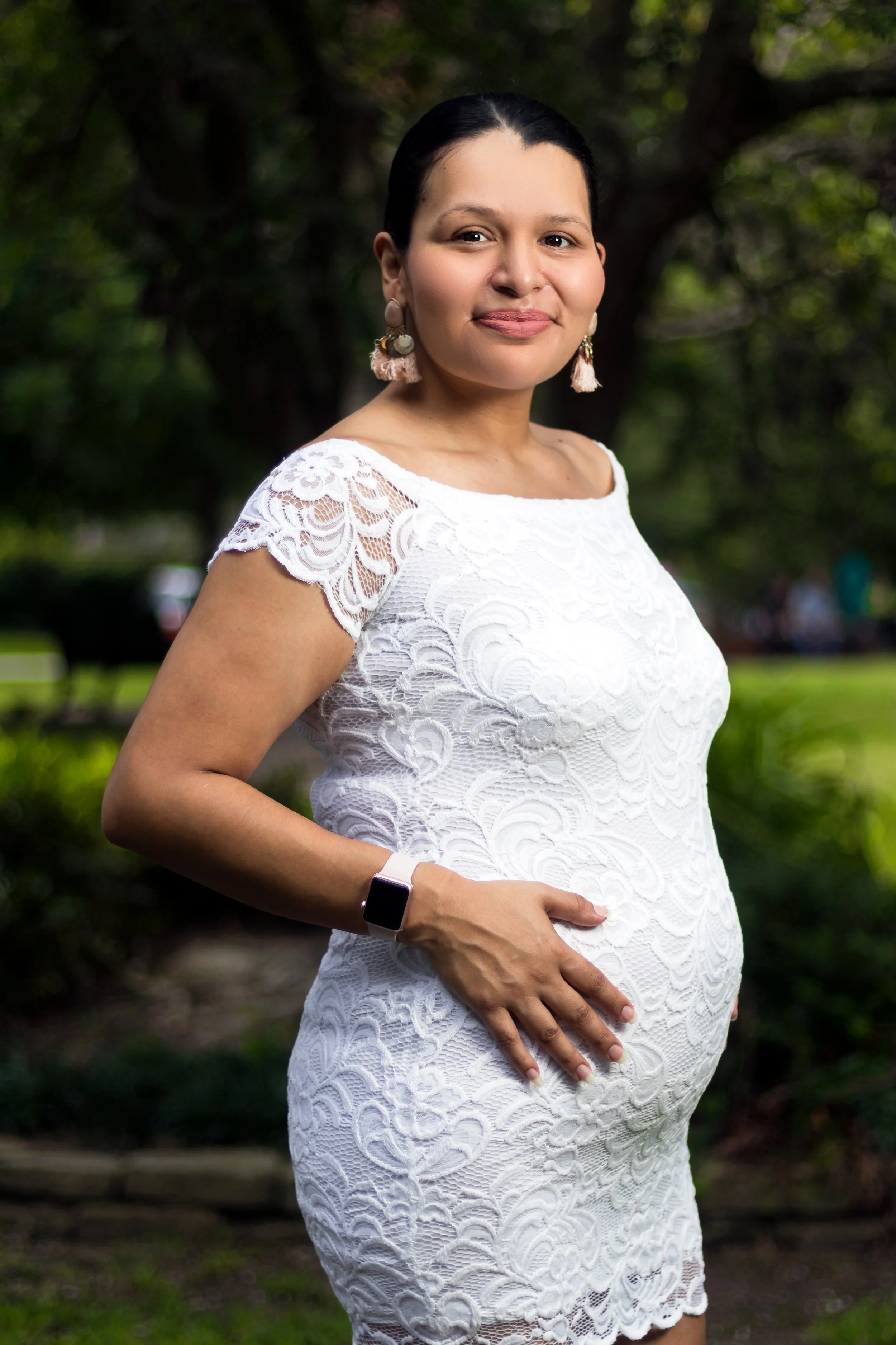 A pregnant woman in a white lace dress standing outdoors in a park, smiling gently, with one hand on her belly and the other at her side. She is wearing tassel earrings and a smartwatch.