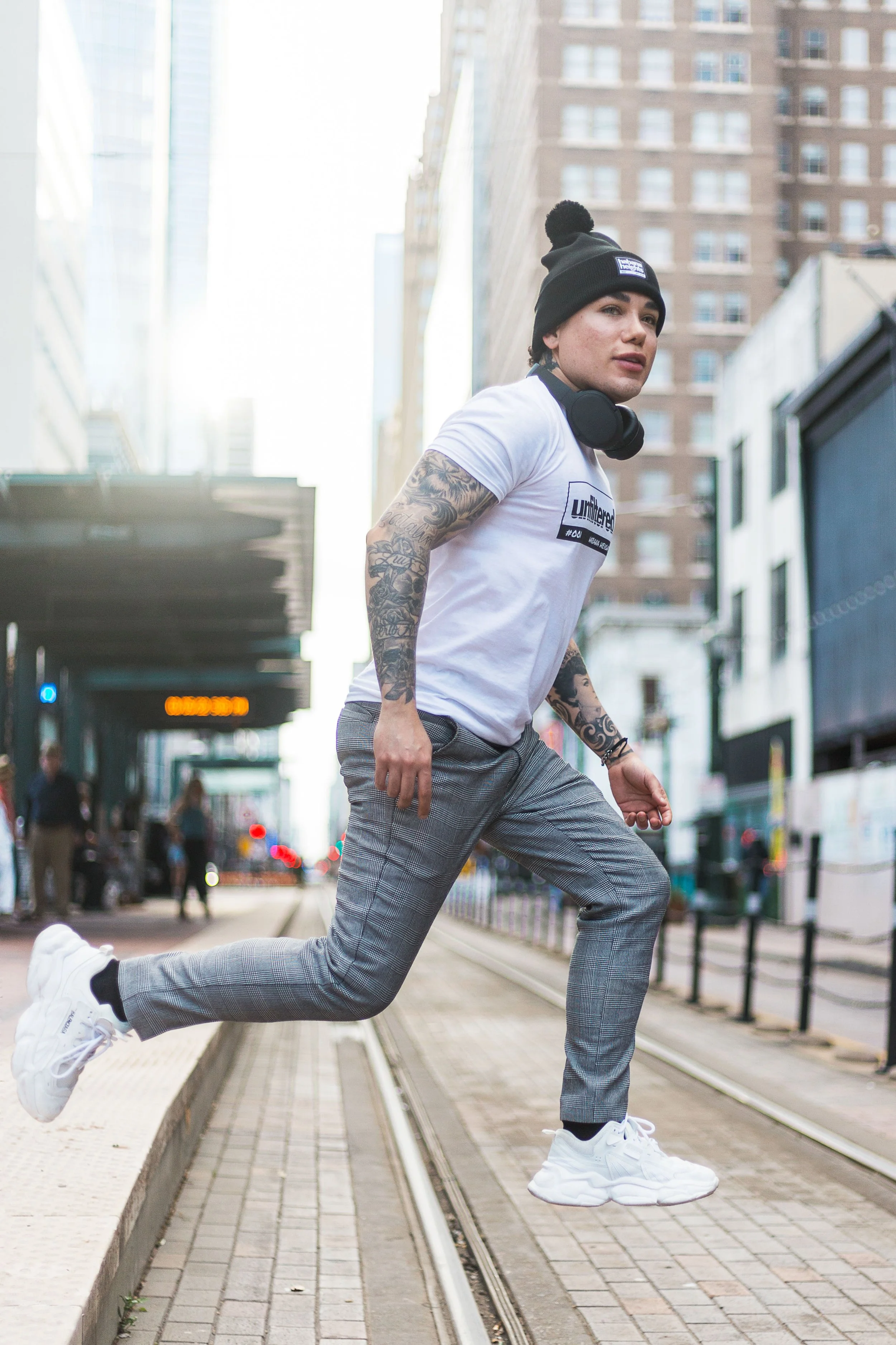 Person jumping on city street with buildings and tram tracks, wearing a black beanie, white t-shirt, checkered pants, and white sneakers.