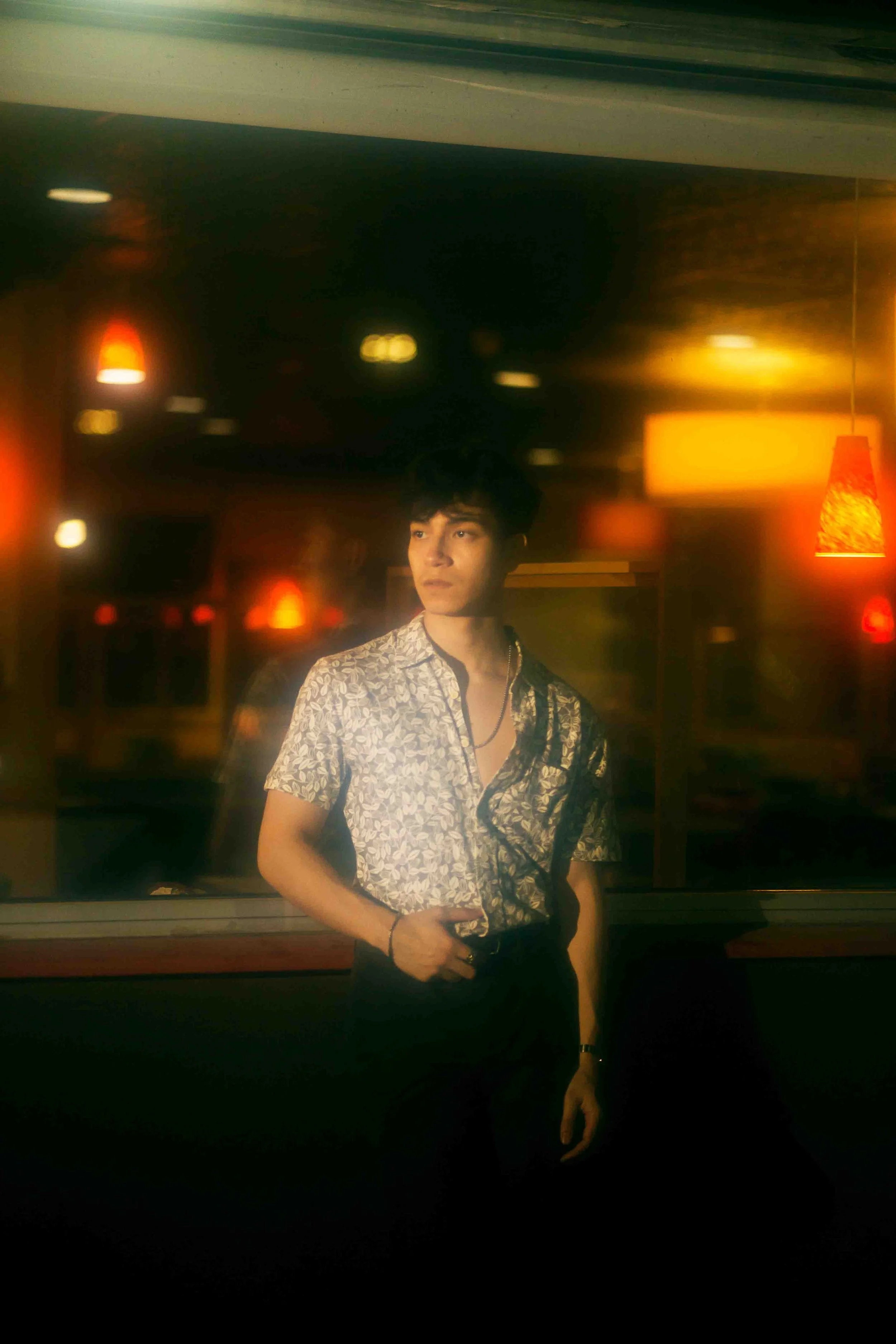Houston editorial portrait of a young man with dark hair wearing a patterned shirt and black pants, standing indoors at night with warm lighting and hanging lamps in the background