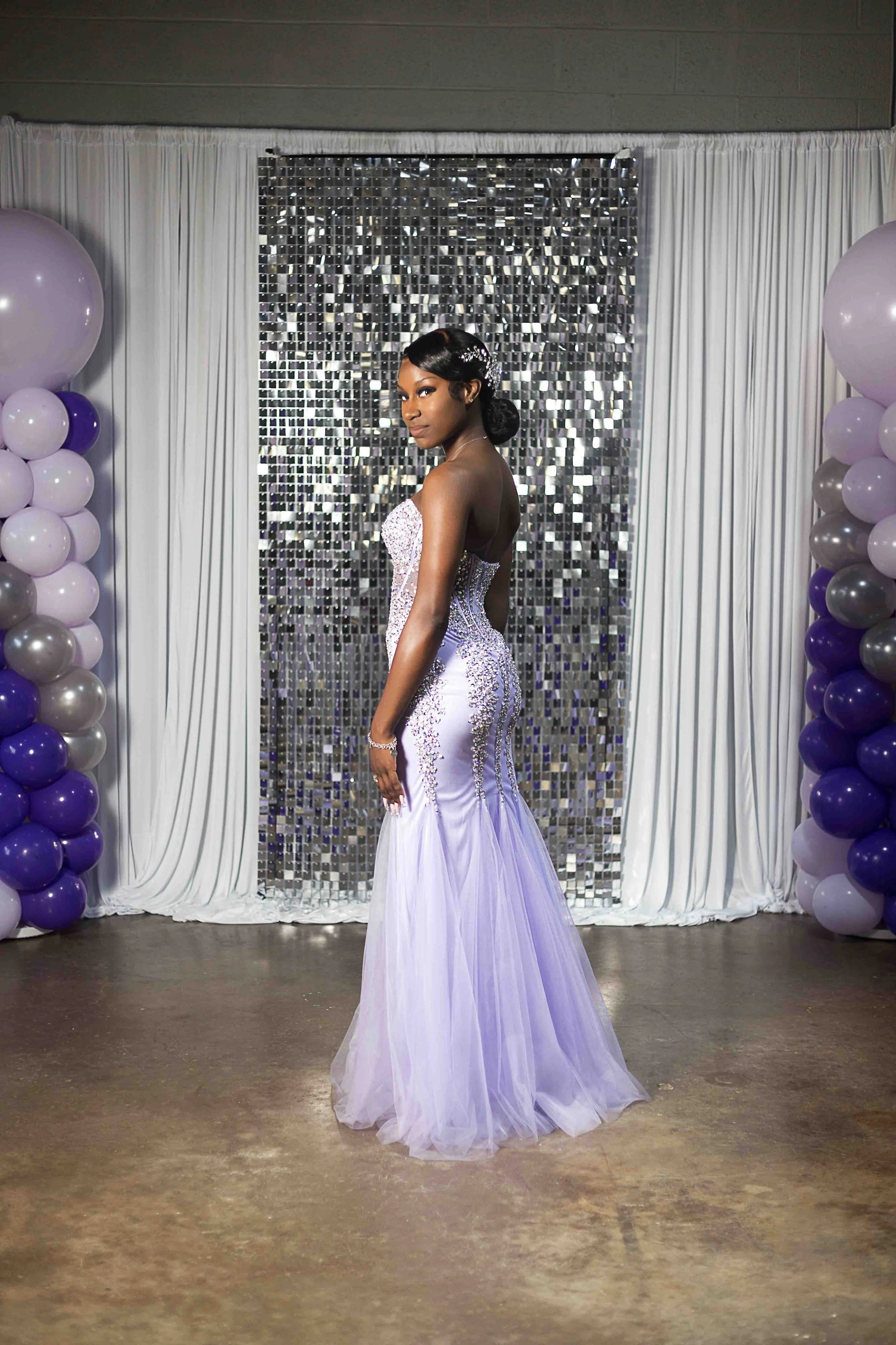Prom portrait of a young woman in a strapless, jewelry-embellished lavender gown standing in front of a silver mosaic backdrop, surrounded by purple and white balloons and white drapery