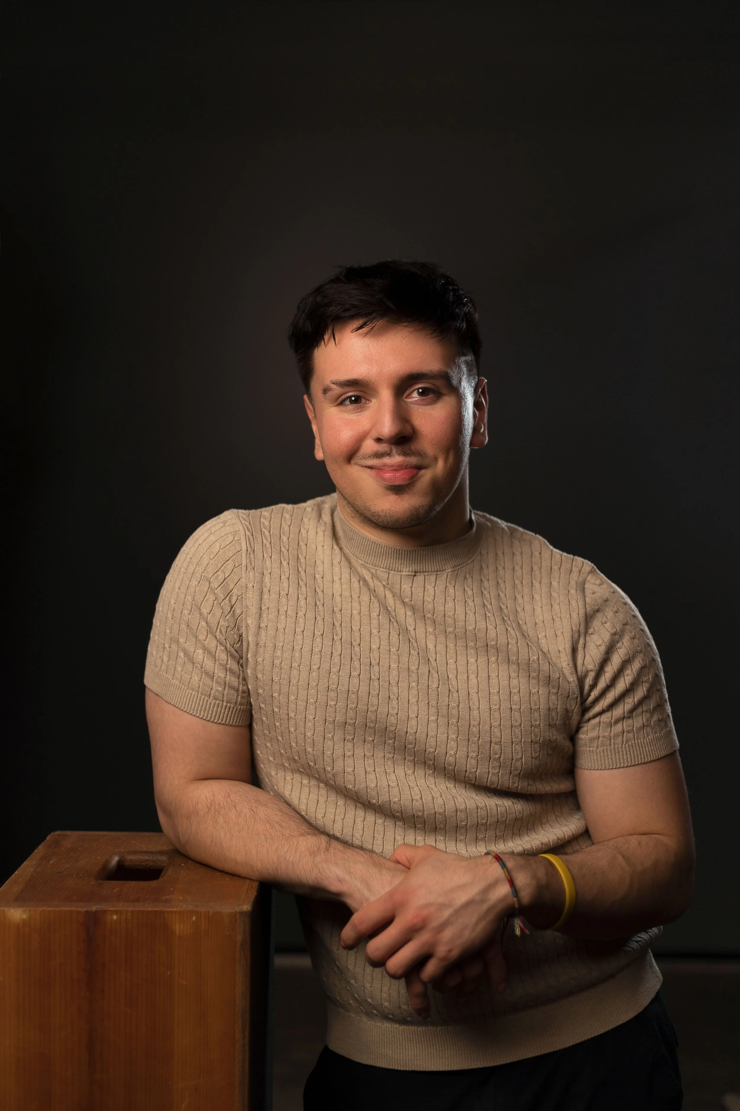 A young man with dark hair and a light skin tone, wearing a beige cable-knit t-shirt, sitting on a wooden block against a dark background, smiling softly at the camera.
