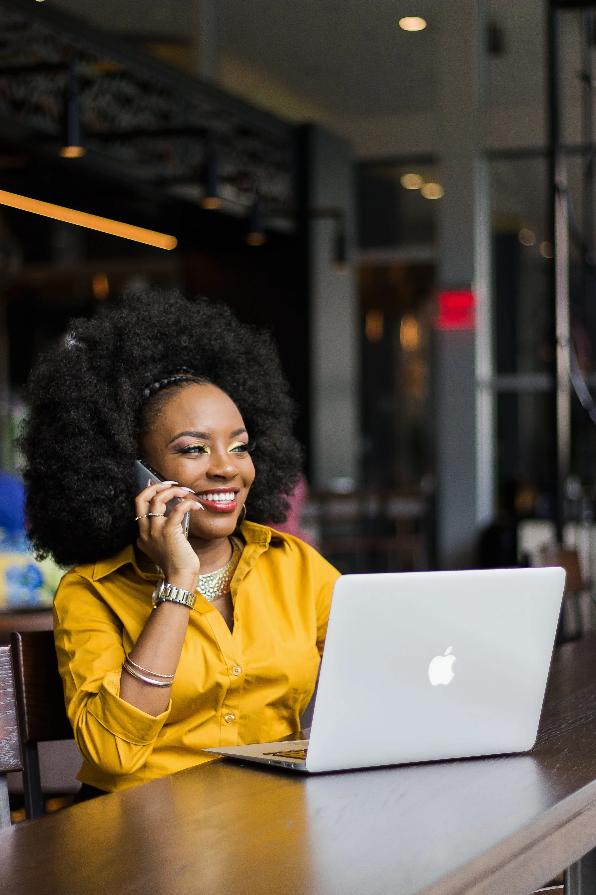 A woman with curly hair, wearing a yellow blouse and jewelry, is sitting at a table in a cafe or restaurant, talking on the phone, with an open MacBook in front of her.
