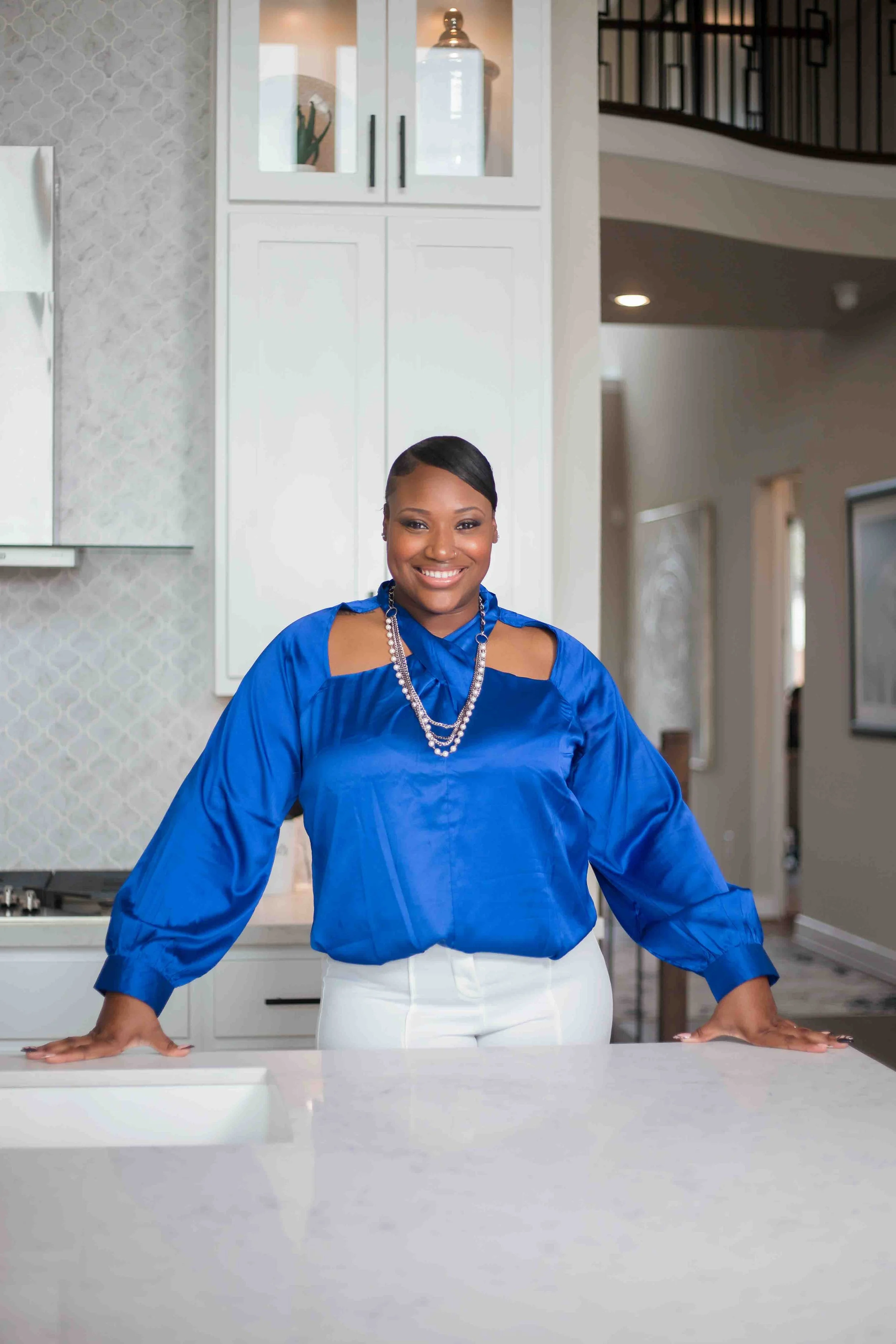 Katy, Texas real estate photography by Lev's Photography featuring a professional female realtor in a bright blue blouse and white pants standing in a modern kitchen, smiling confidently for realtor branding and marketing photos, ideal for real estat