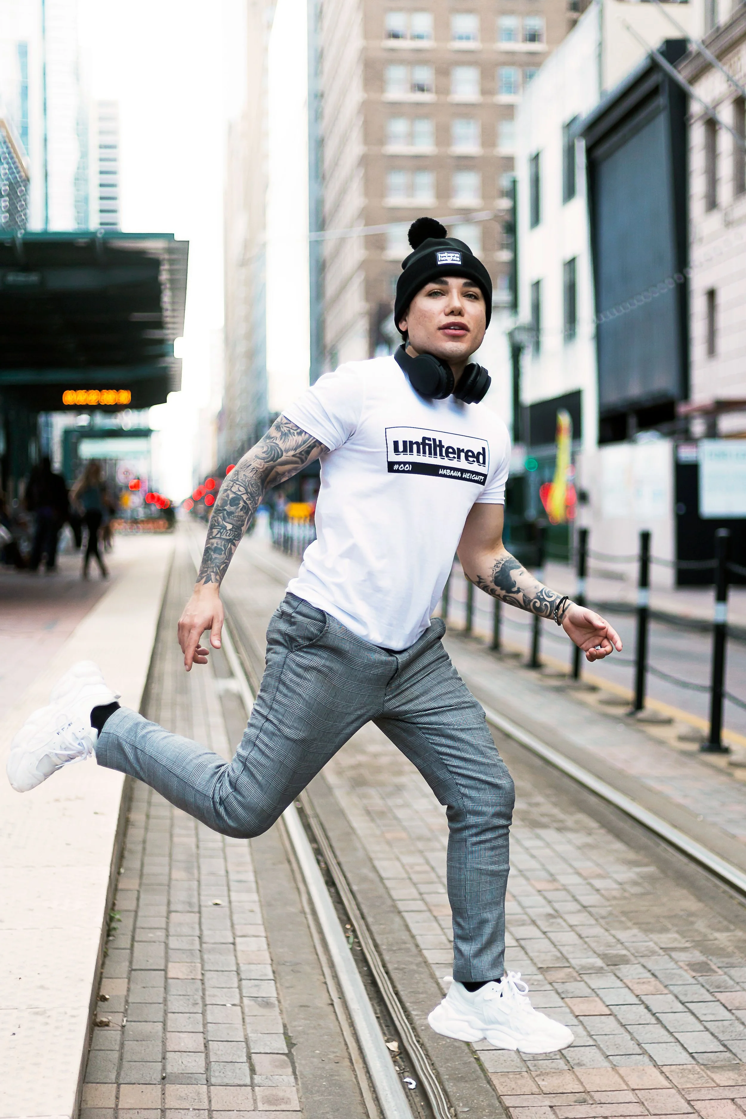 A young woman with tattoos on her arms, wearing a black beanie, white t-shirt with 'unfiltered' printed on it, gray checkered pants, and white sneakers, is running on city street tracks surrounded by tall buildings.