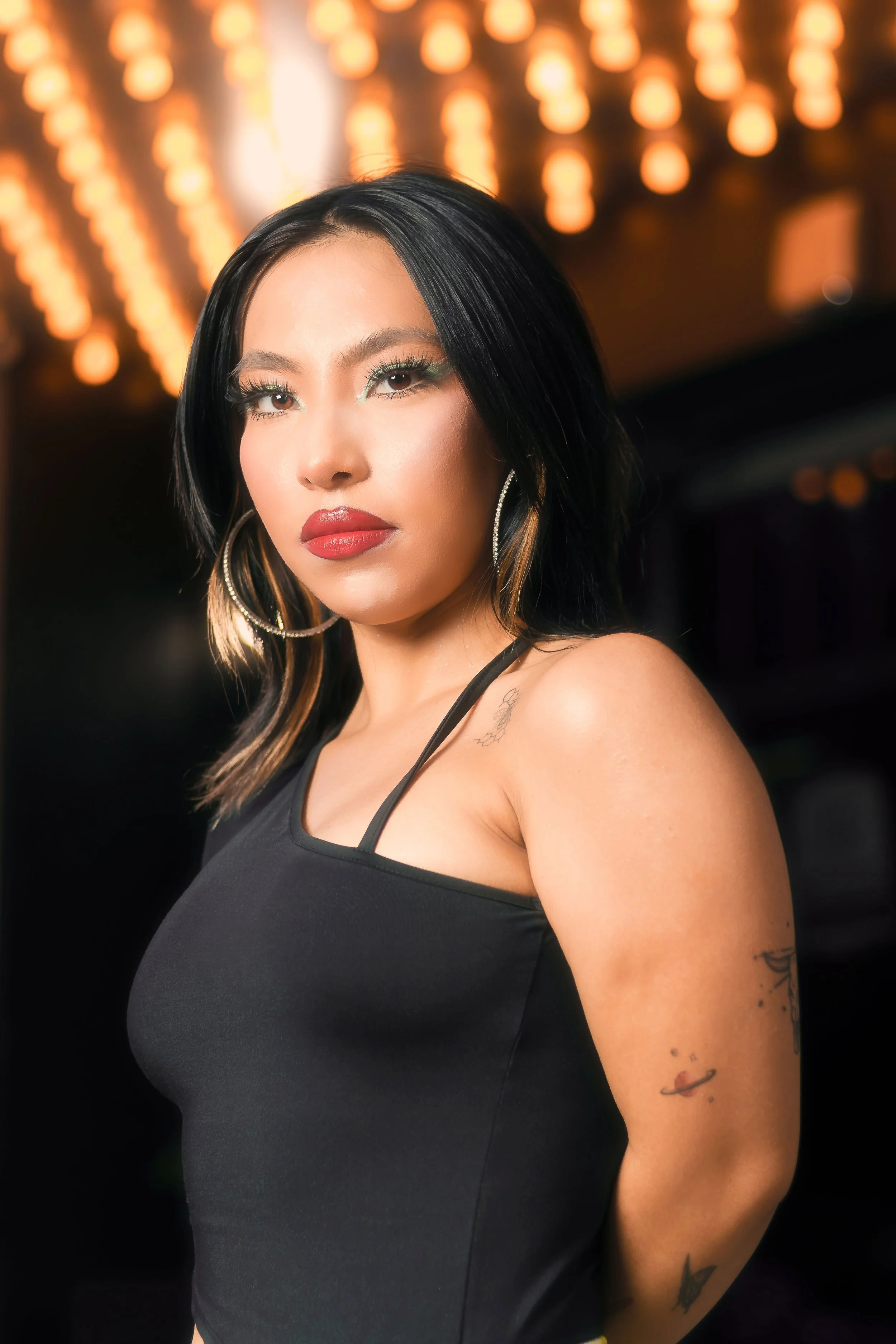A woman with black hair, bold makeup, hoop earrings, and tattoos, wearing a black sleeveless top, standing in front of a background with blurred orange lights.