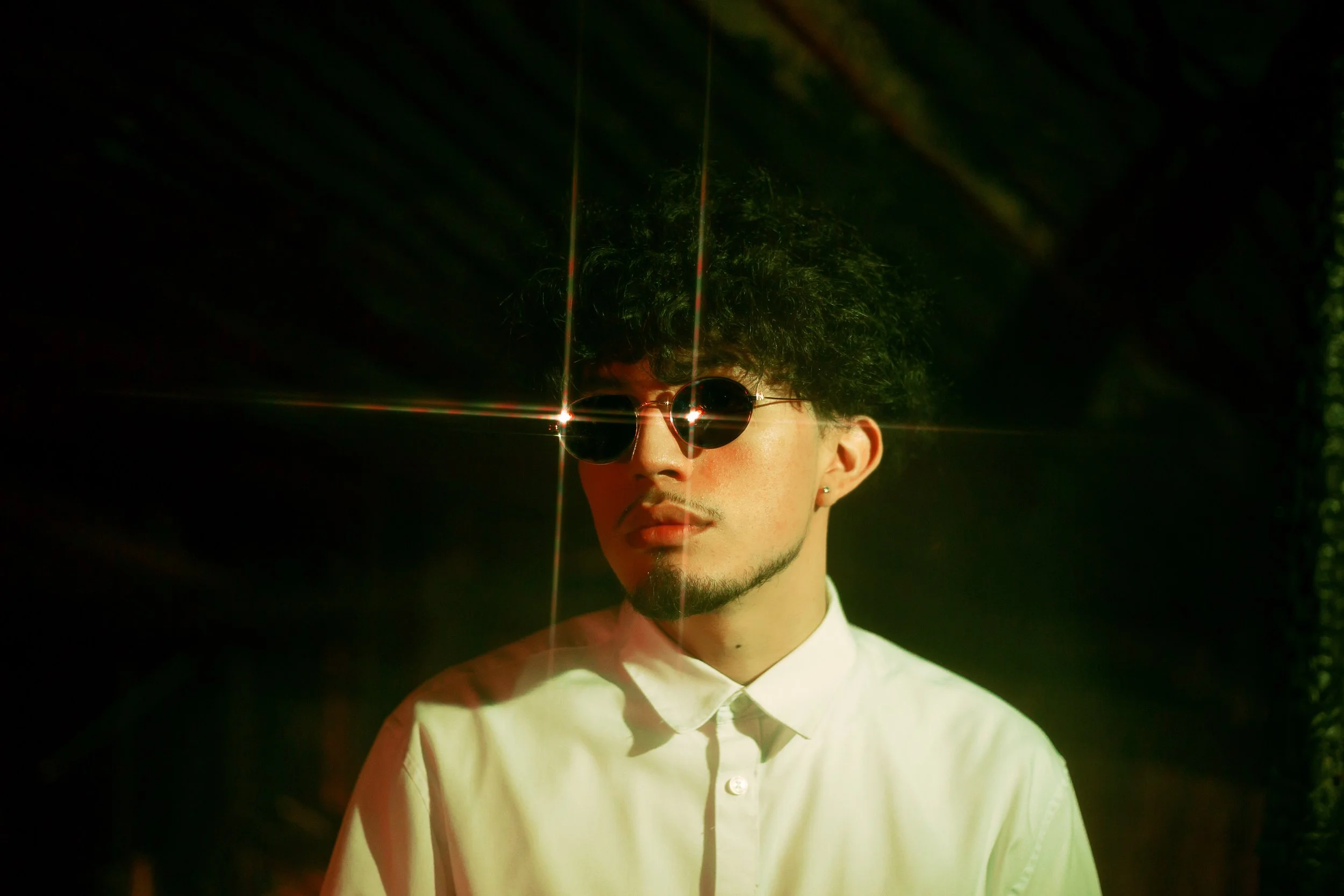 A young man with curly hair, wearing sunglasses and a white button-up shirt, standing in a dark environment with a lens flare effect creating bright star-like reflections around his face.
