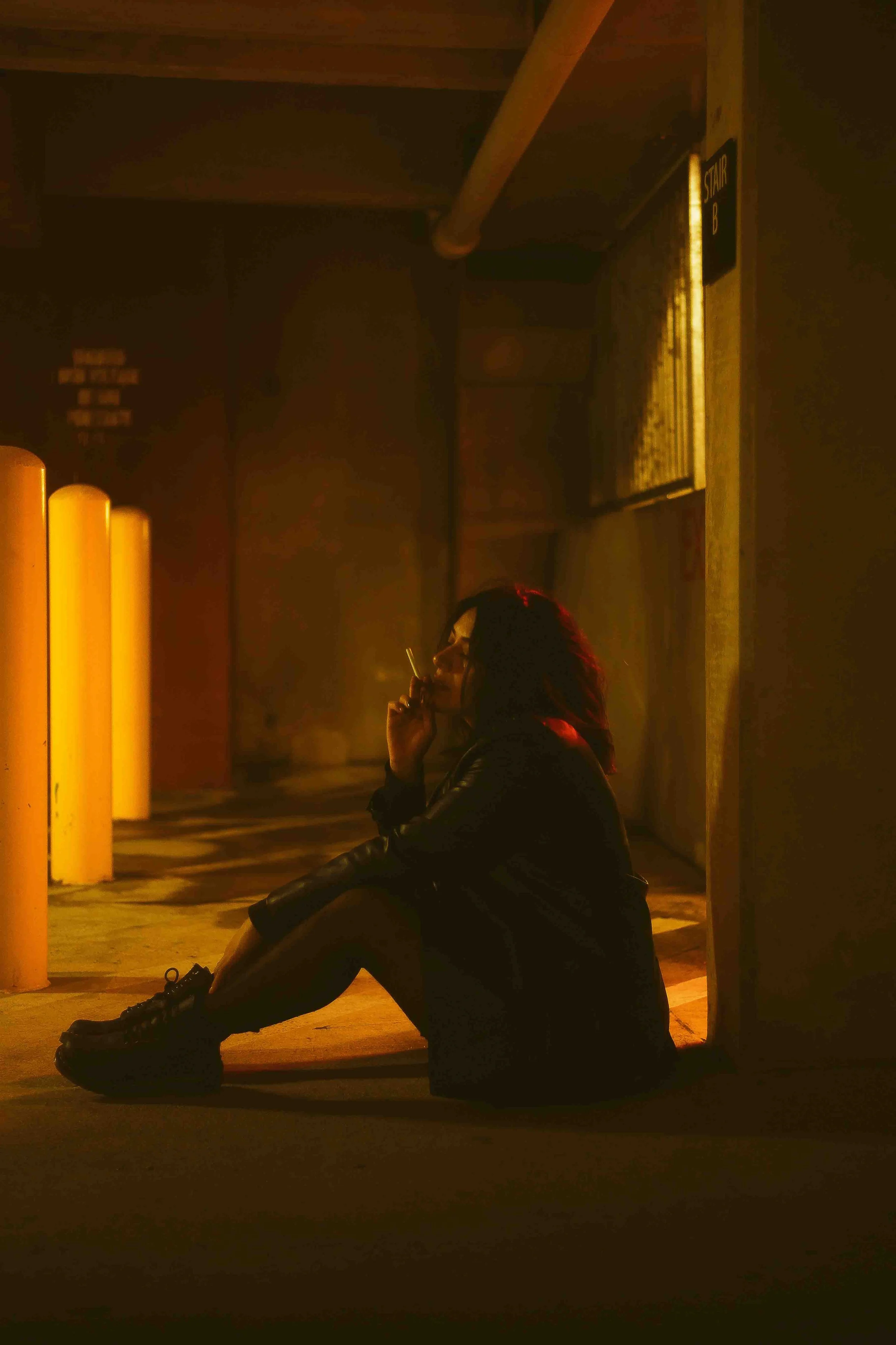 Cinematic night portrait of a woman sitting on the ground outside a building entrance, smoking a cigarette under warm yellow-orange lighting