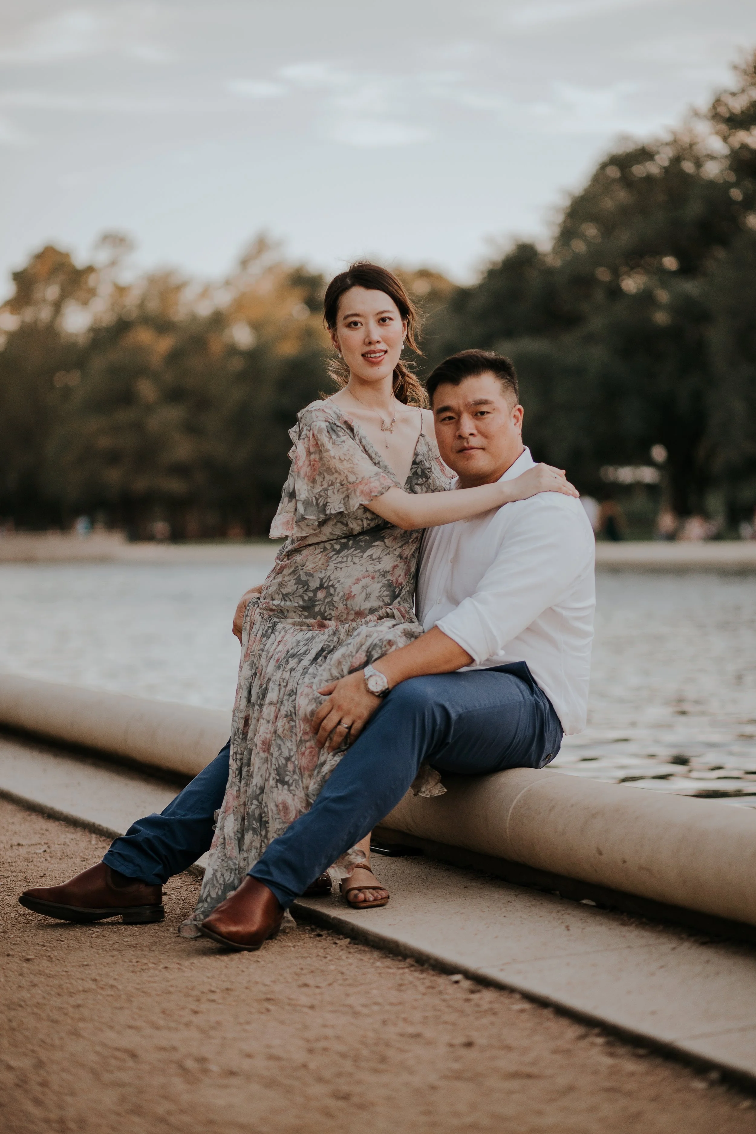 A couple sitting by a river, with the woman on the man's lap, during sunset in a park. Both are dressed casually, and the background shows trees and water.