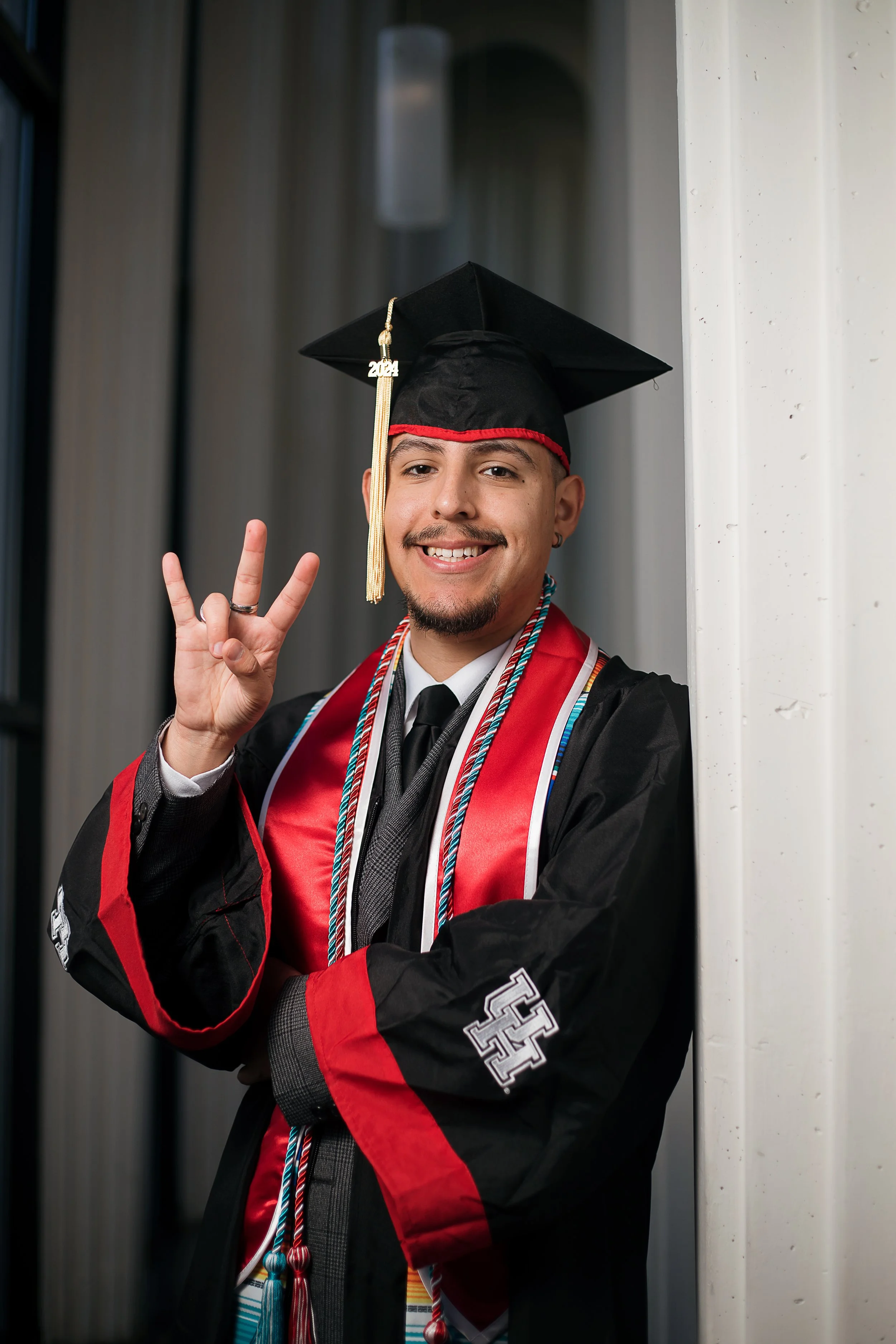 A young man in a graduation cap and gown smiling and making a peace sign with his right hand at a graduation ceremony.