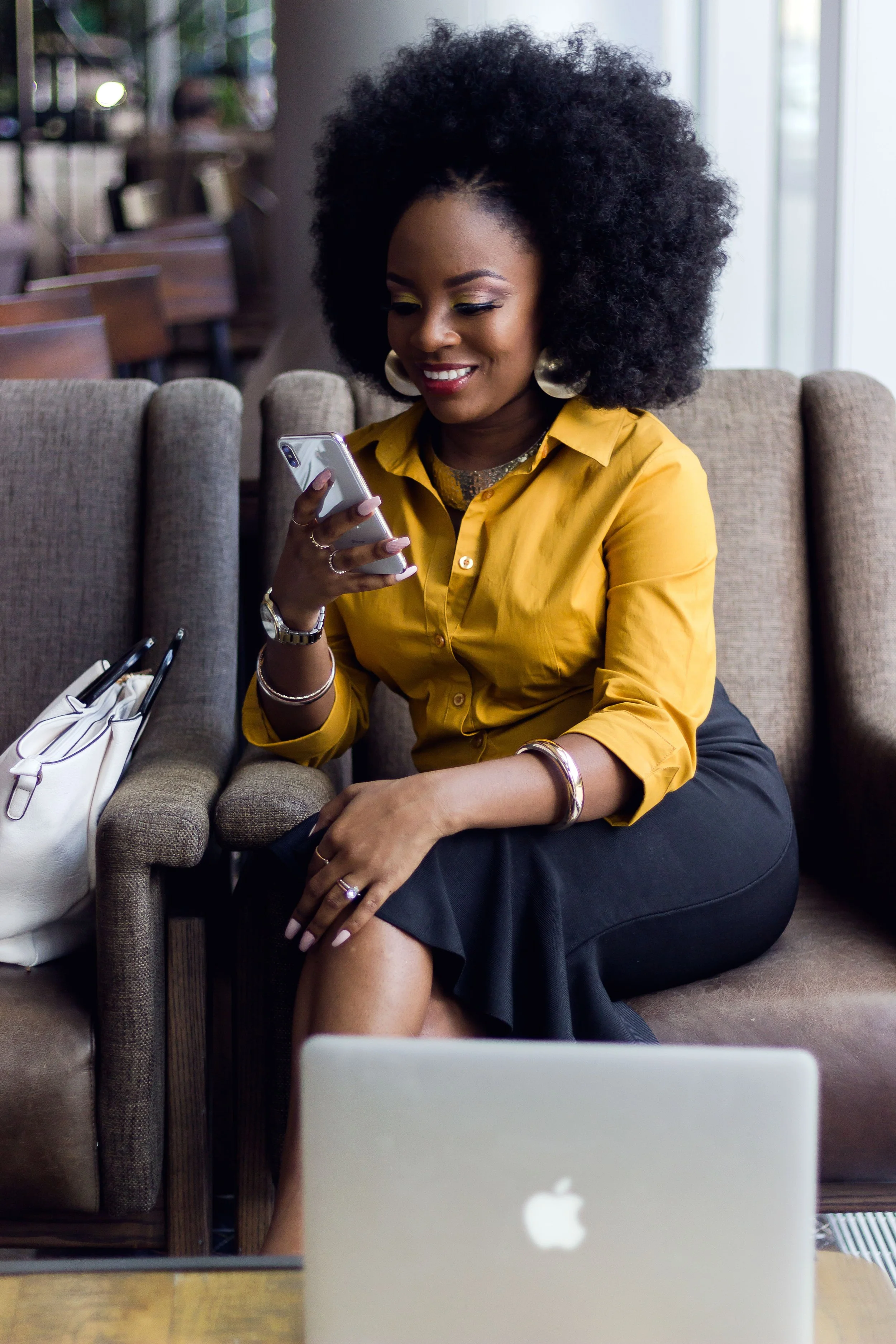 A woman with curly black hair and gold earrings sitting on a beige couch, smiling and looking at her phone, with a silver MacBook on the table in front of her.