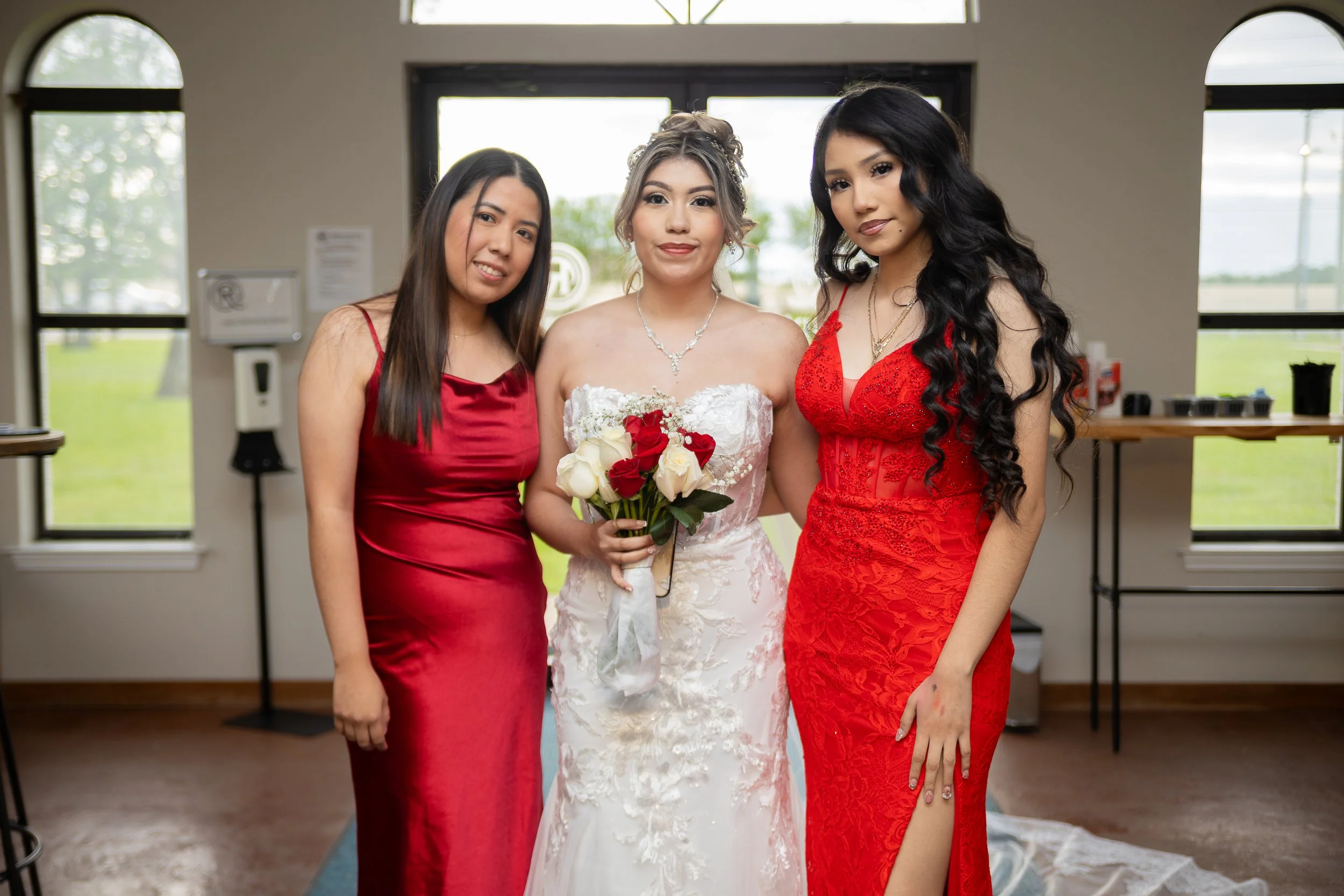 Three women in formal dresses standing indoors, with a bride holding a bouquet of red and white roses.