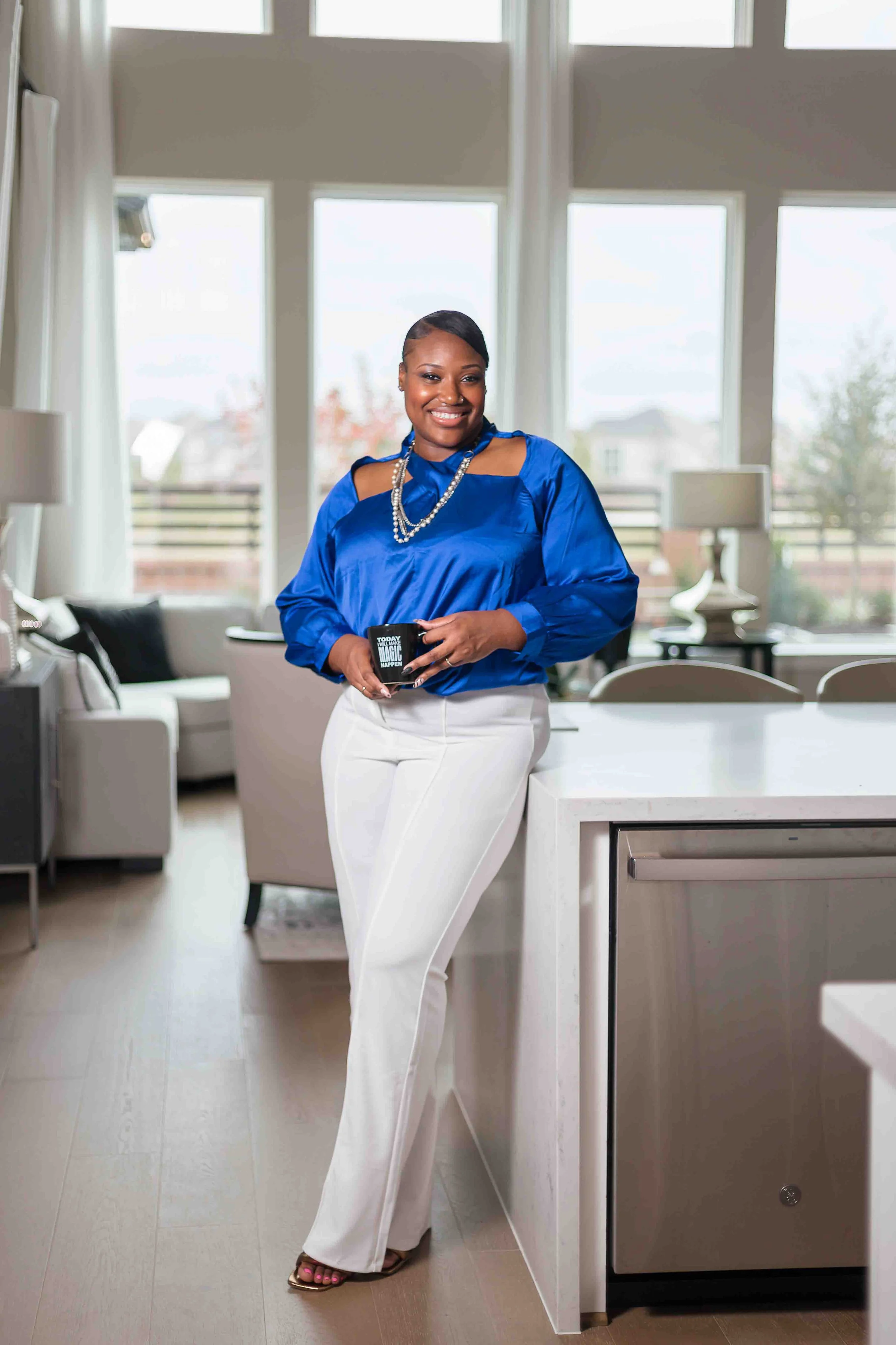 Katy, Texas realtor branding portrait by Lev's Photography featuring a professional woman standing in a bright modern kitchen, holding a coffee mug, smiling, wearing a blue top and white pants, ideal for real estate social media, marketing, and websi