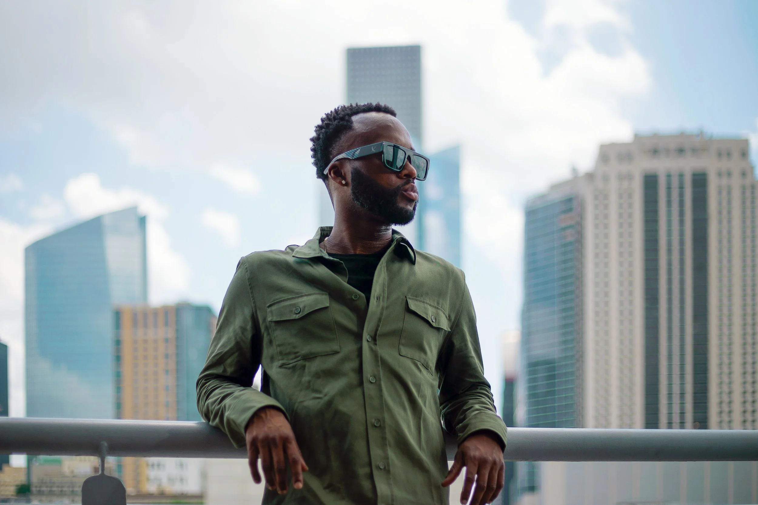 A man wearing sunglasses and a green shirt leans on a railing with city skyscrapers in the background.