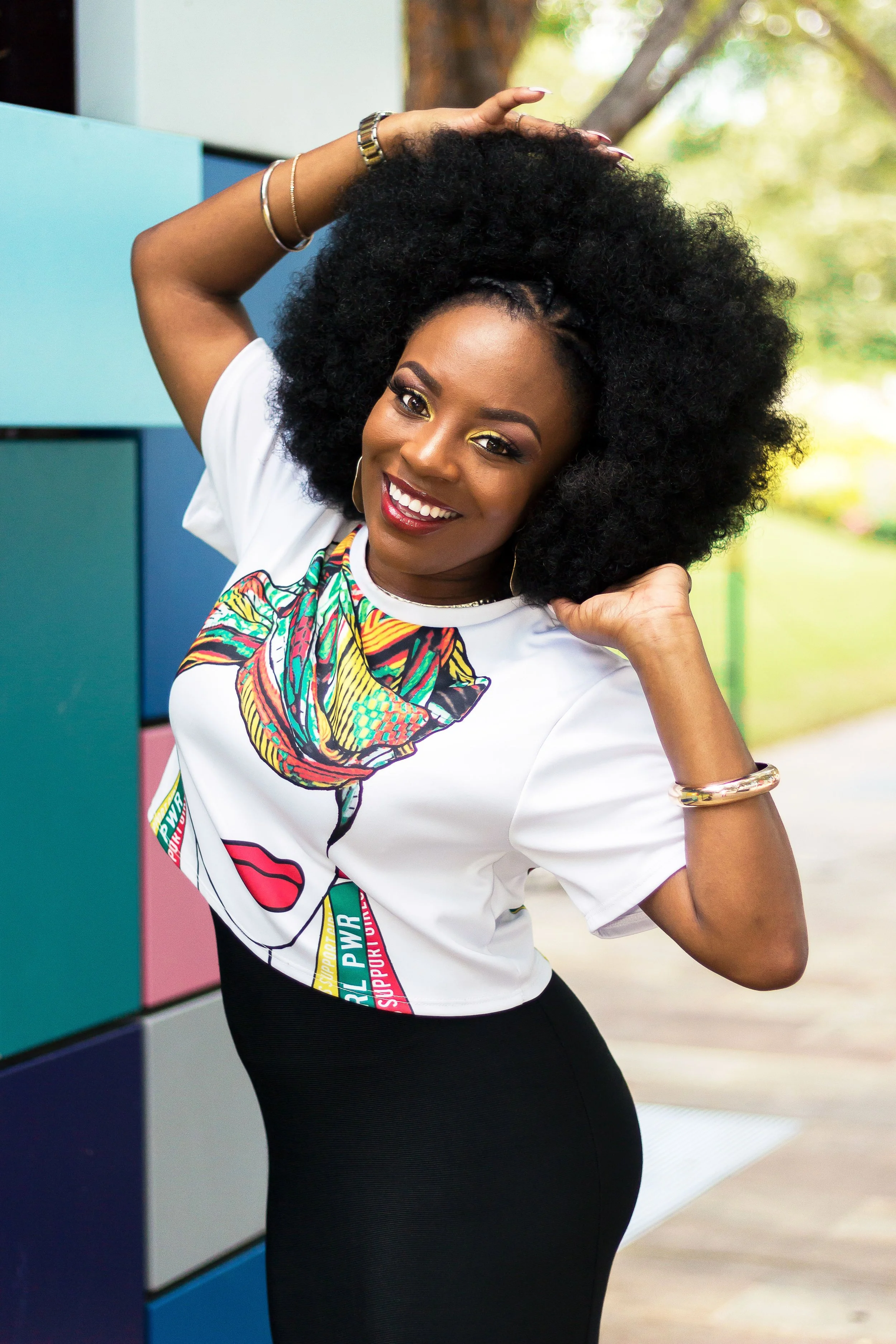 A woman with medium-dark skin, curly black hair, and bold makeup, smiling and posing outdoors. She is wearing a white t-shirt with a colorful, patterned design and the words "Support Girl Power," along with a black skirt and gold jewelry.