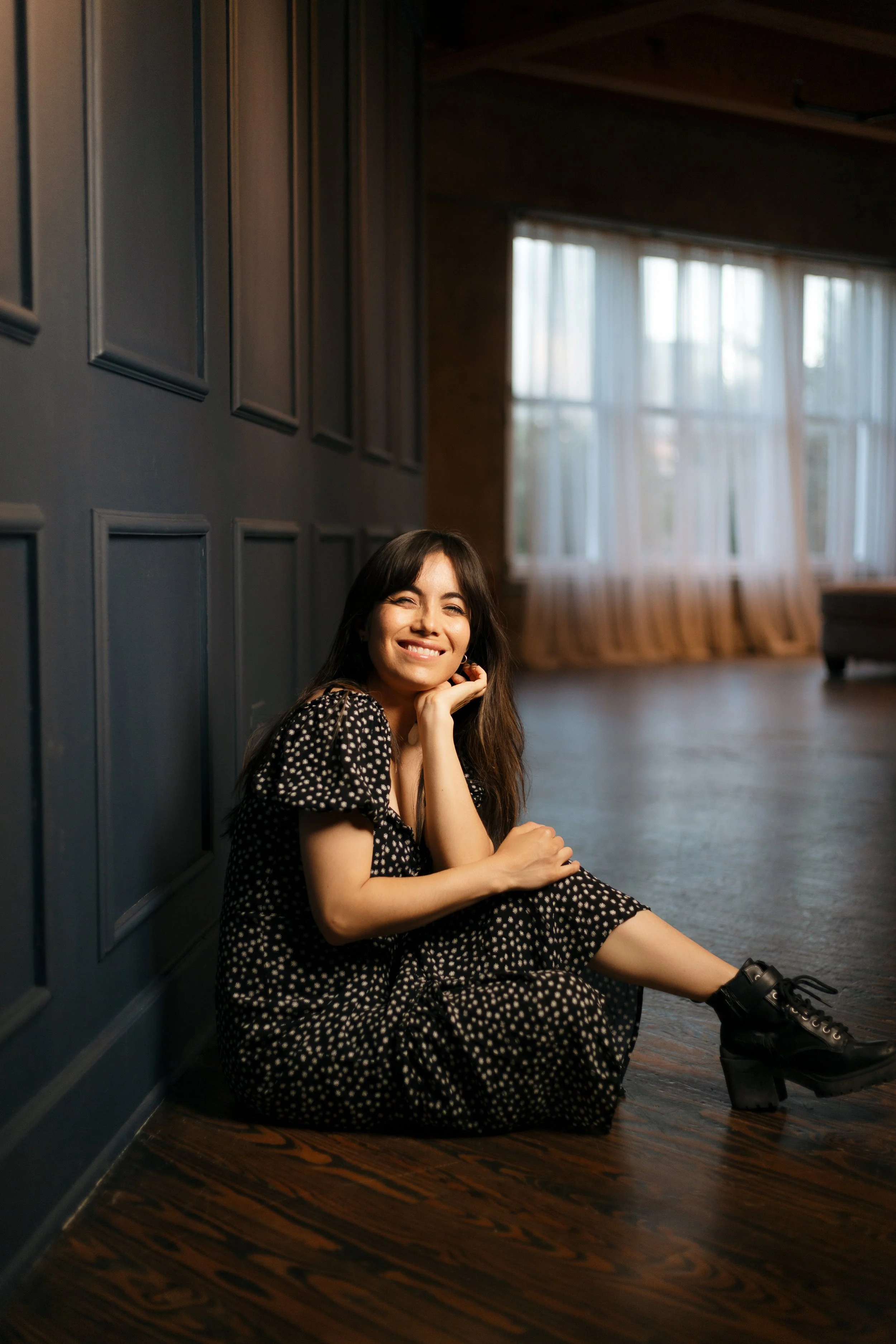 A young woman in a black and white polka dot dress sitting on a hardwood floor, smiling and resting her chin on her hand, in a room with large windows and blue paneled walls.