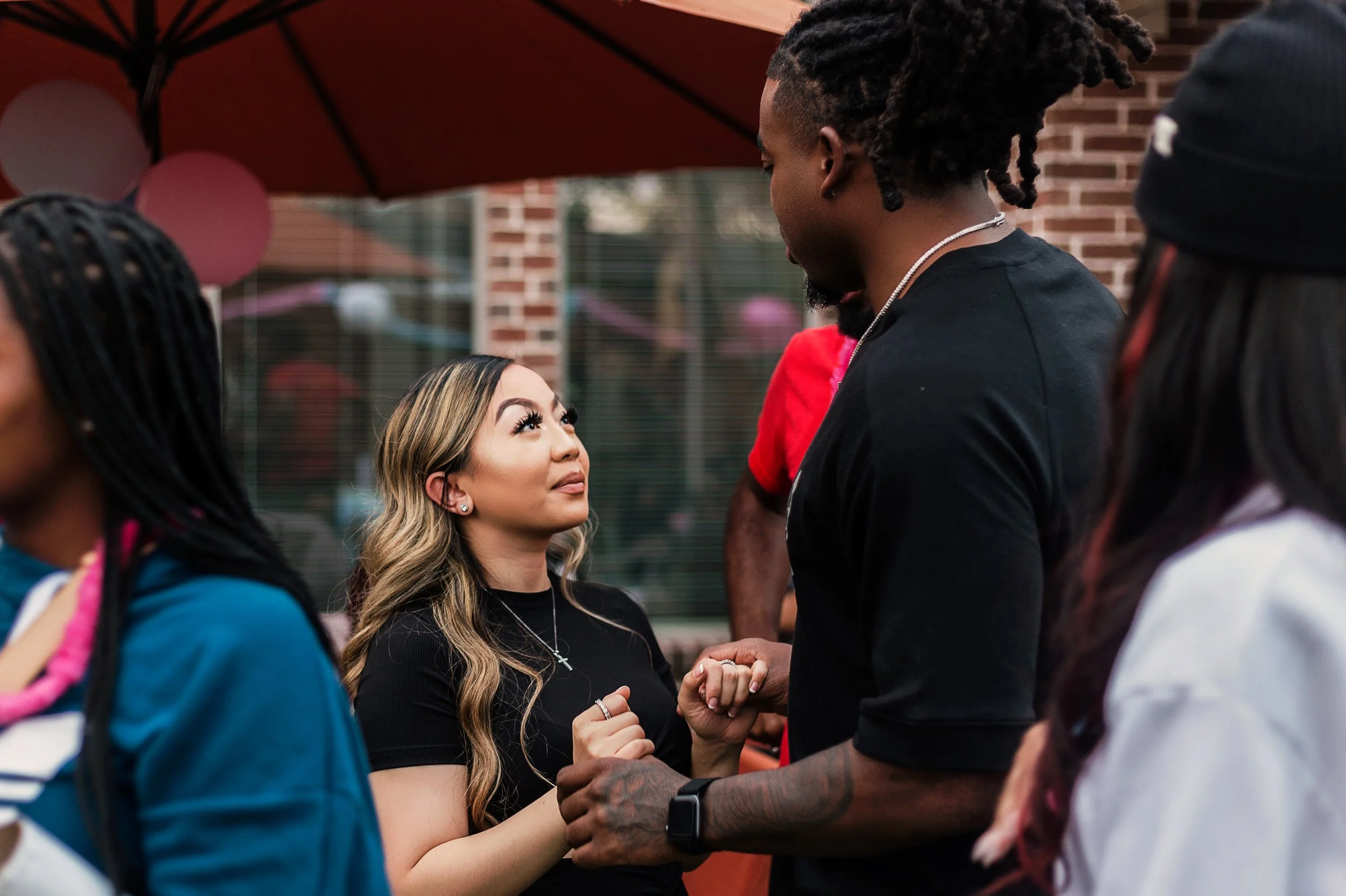 A woman and man holding hands and looking into each other's eyes at an outdoor gathering, with other people and decorations in the background.