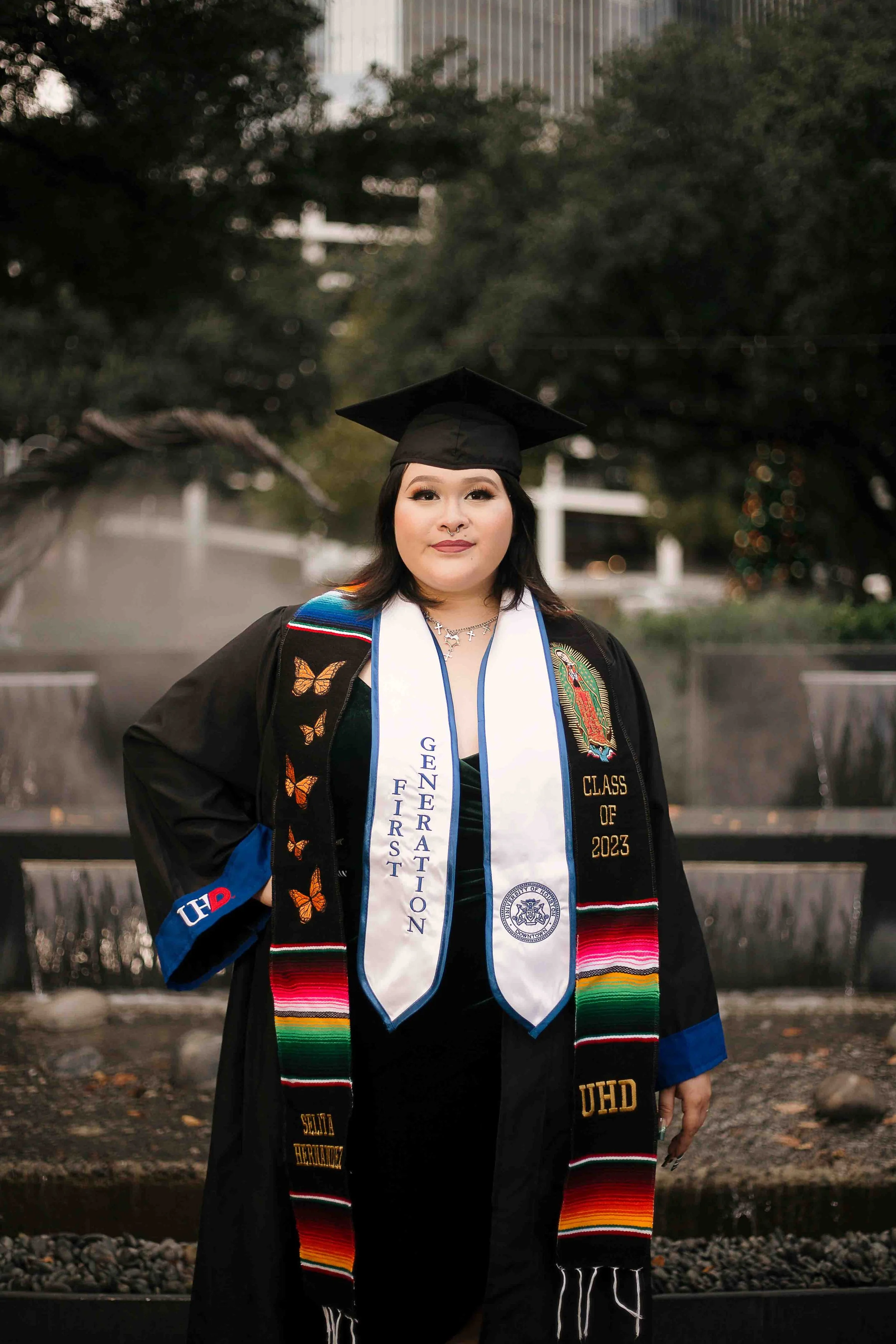 University of Houston–Downtown graduate standing in front of a fountain wearing a cap and gown with a decorated stole and a white sash reading “Genuine First Generation,” celebrating her 2023 graduation