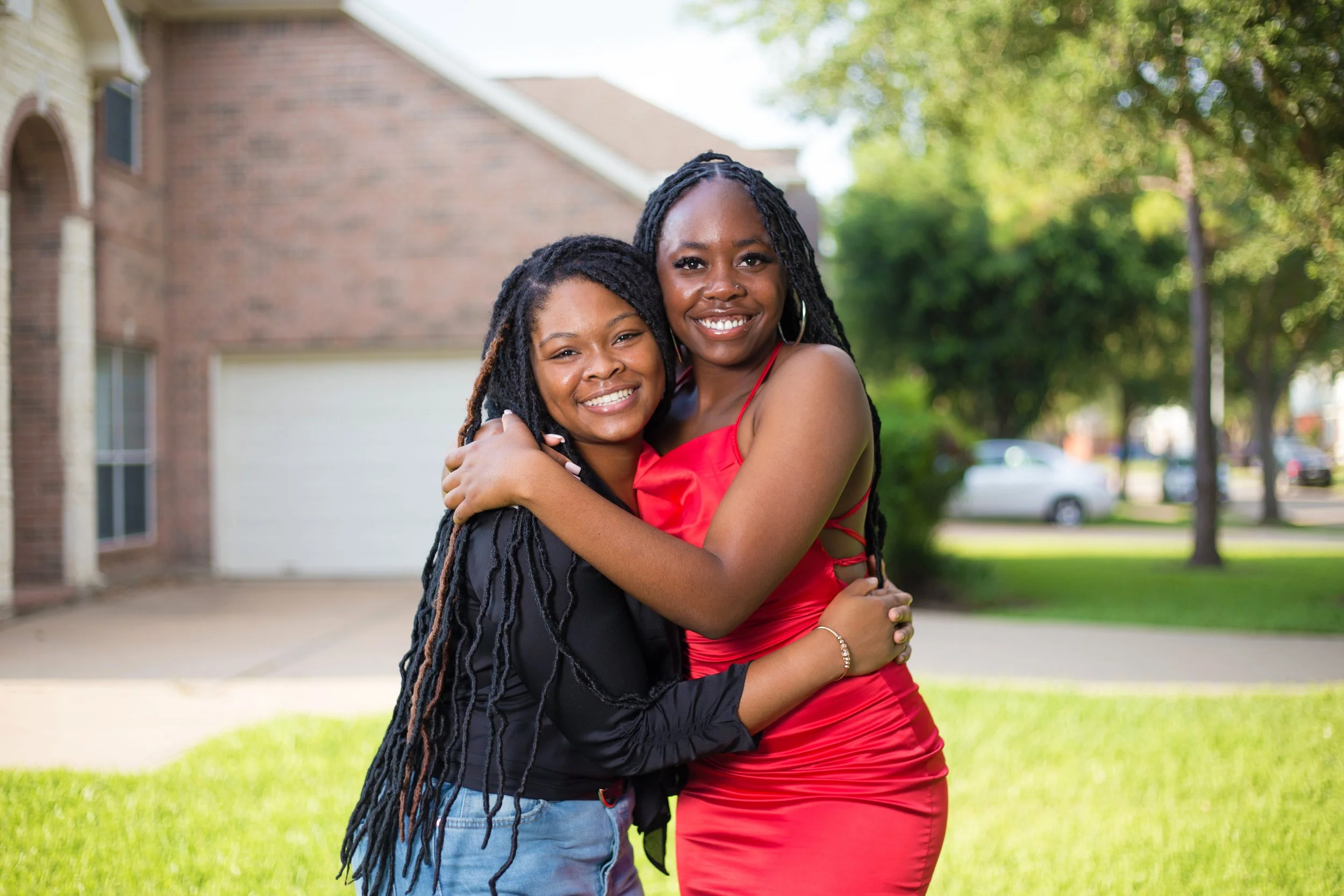 Two women hugging and smiling outdoors in front of a house and trees, one wearing a black jacket and the other in a red dress.